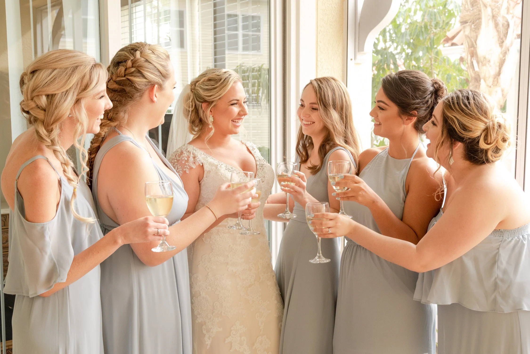 A bride in a wedding dress smiling and holding a glass of champagne, surrounded by six bridesmaids in matching light gray dresses, all holding glasses of champagne and sharing a joyful moment.