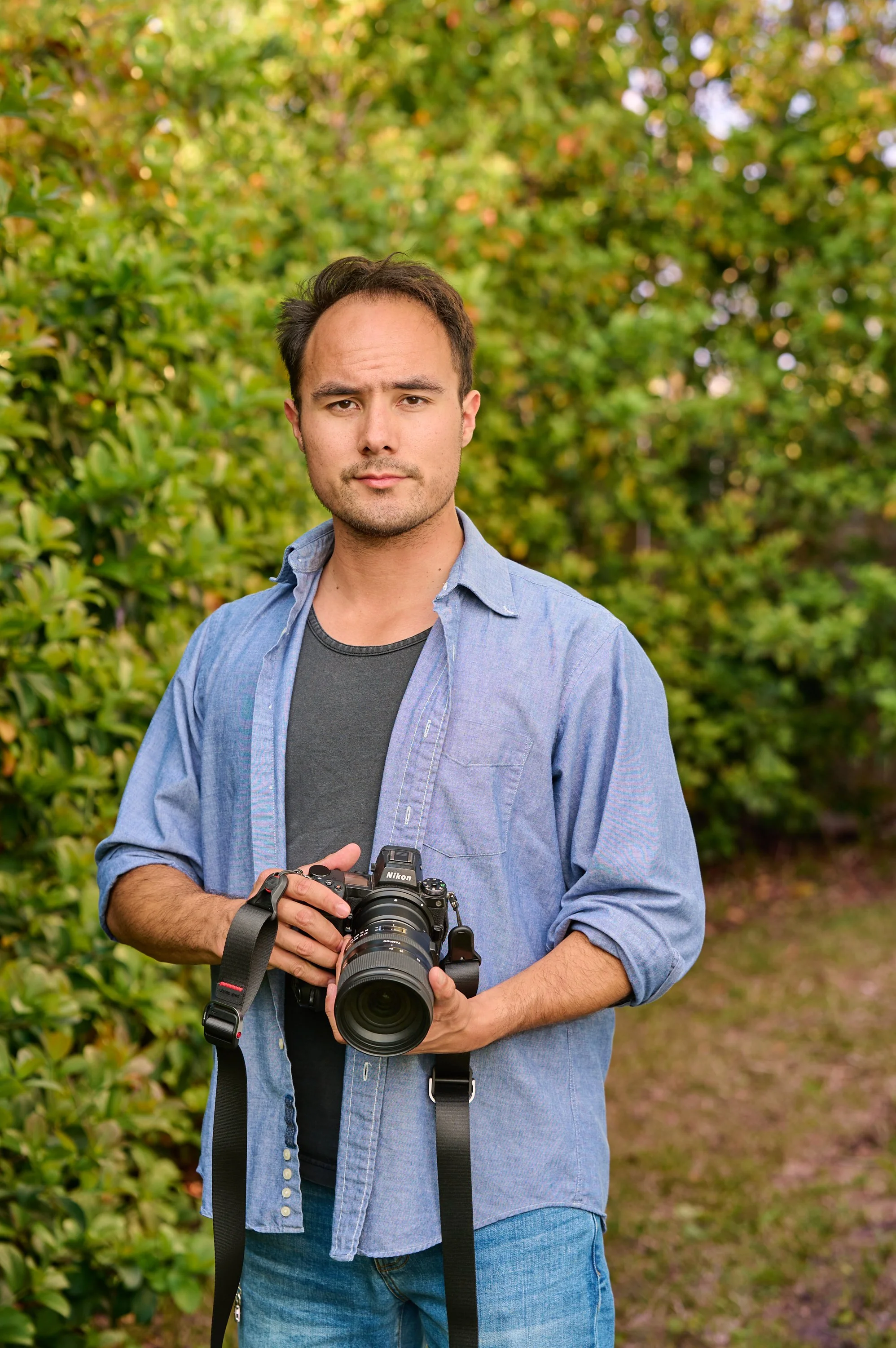 A young man standing outdoors holding a camera, with green foliage in the background.