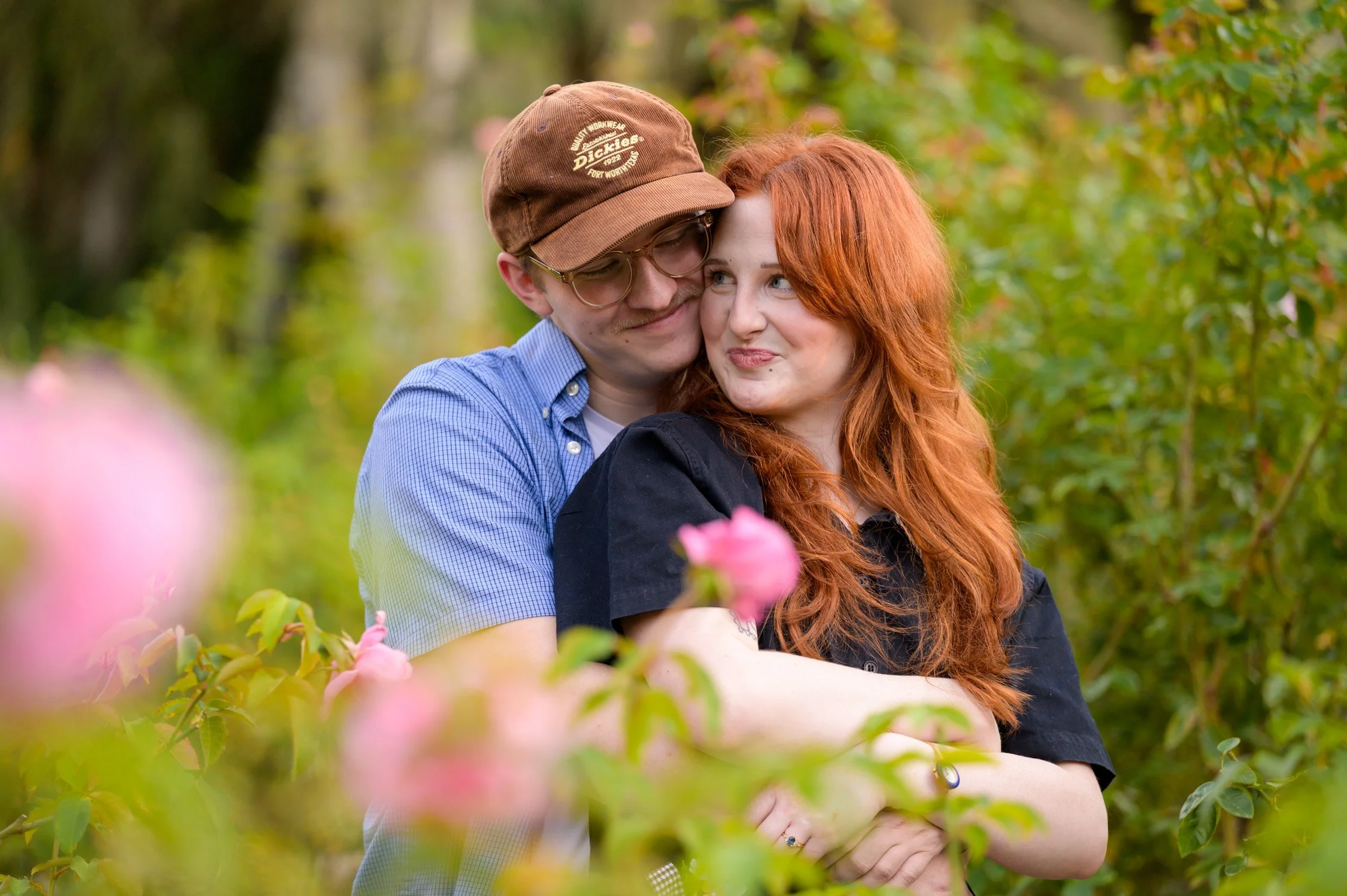 A young couple embracing outdoors among pink flowers and green foliage, smiling and sitting close together.