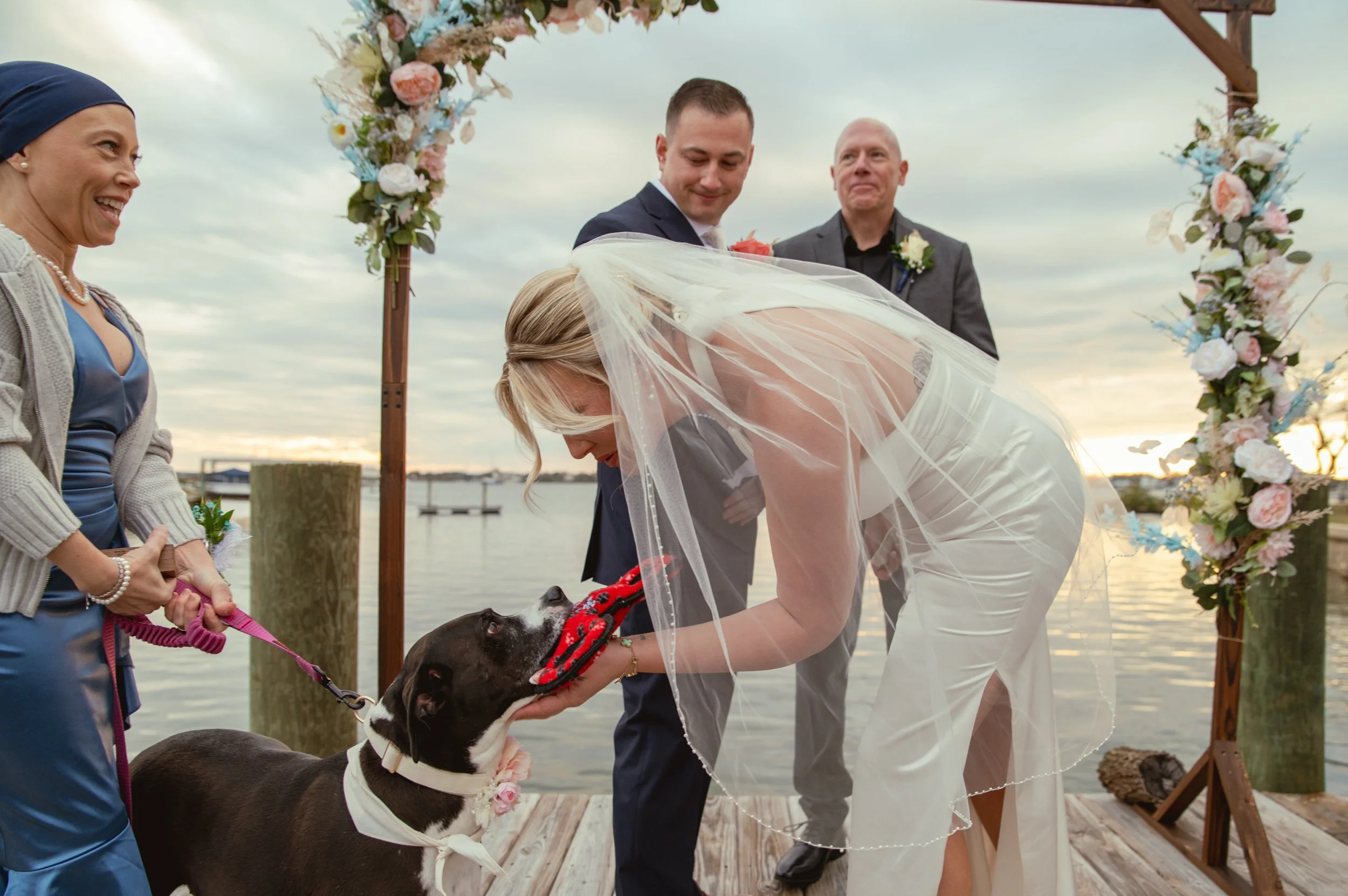 Bride in a white wedding dress leaning forward to take a toy from a dog during outdoor wedding ceremony by water, with groom and officiant, floral arch, and water in background.