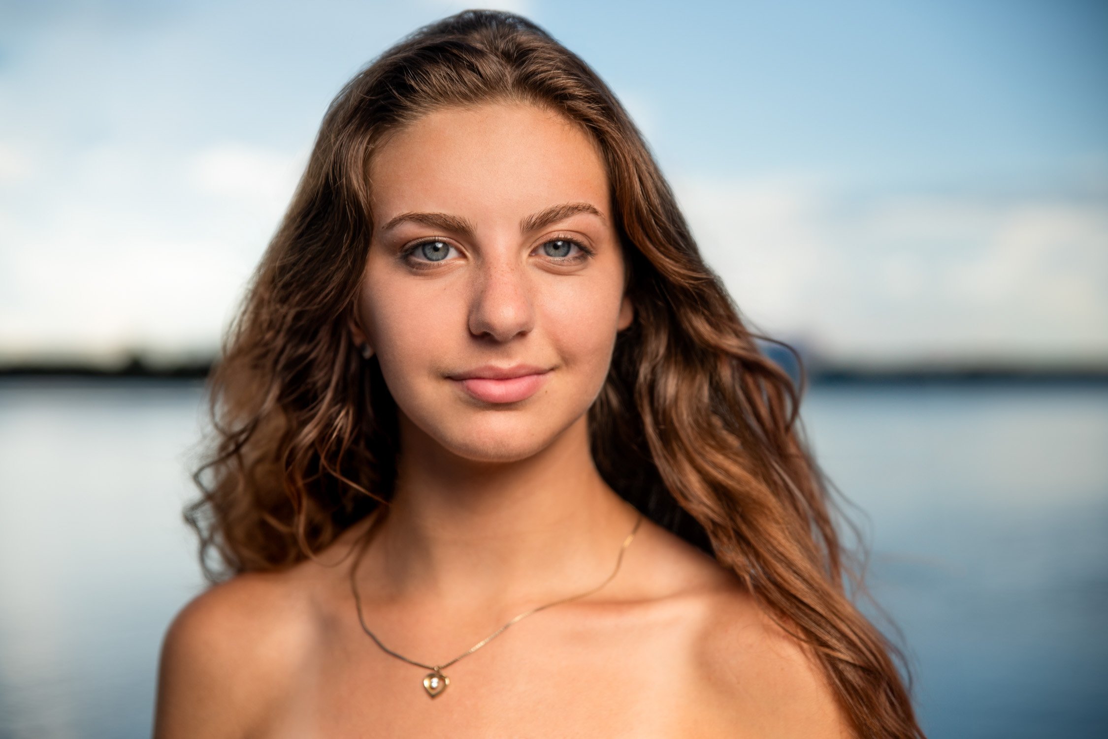 A young woman with long, wavy brown hair and blue eyes is outdoors near a body of water.