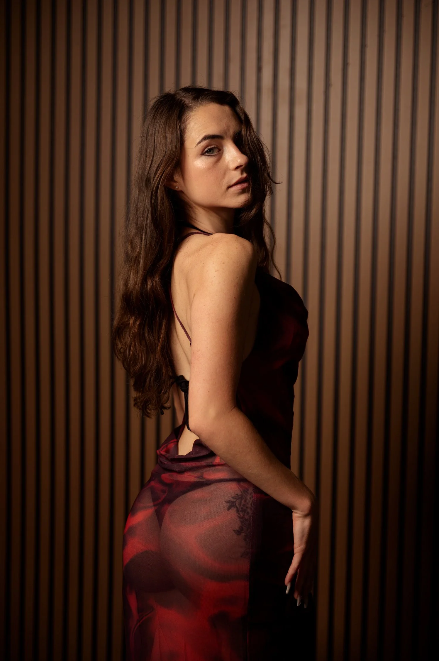 A woman with long brown hair stands against a wooden wall, wearing a dark satin dress with spaghetti straps.