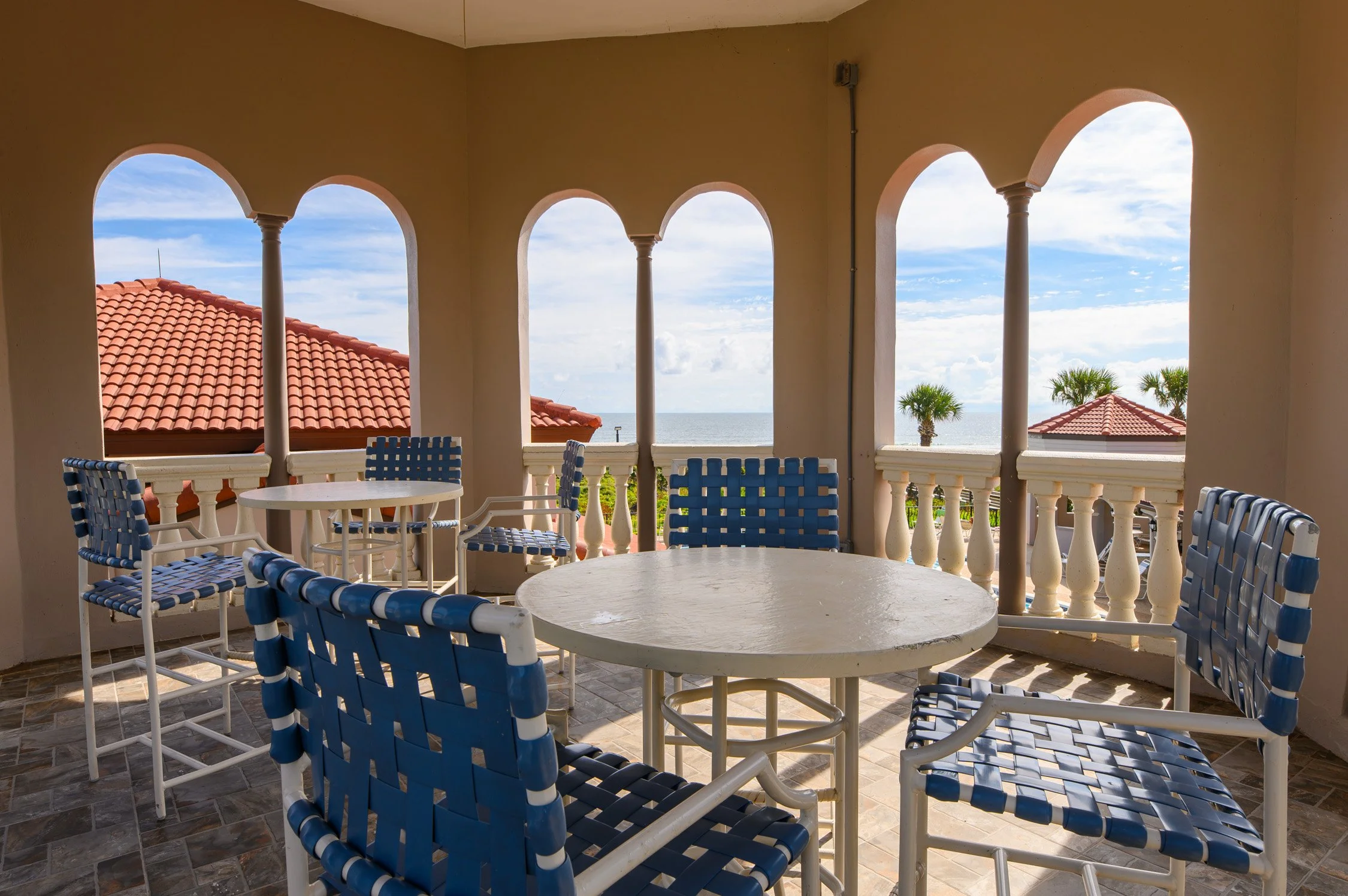 Beachside balcony with three round white tables and six blue woven chairs, arched windows with views of red tile roofs, palm trees, and ocean in the background.