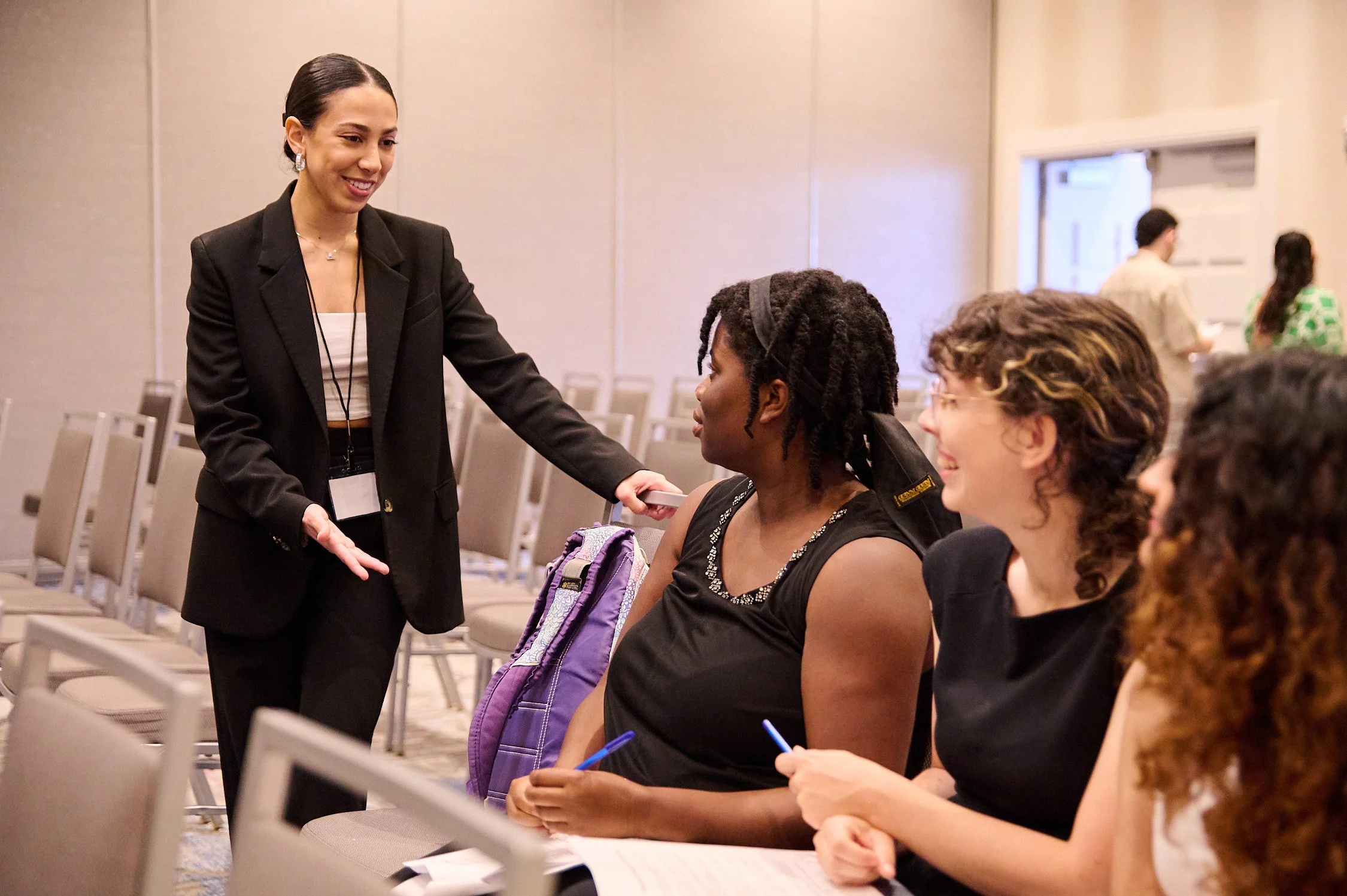 A woman in a black blazer and white top is smiling and talking to three seated women at a conference, with chairs and other people in the background.