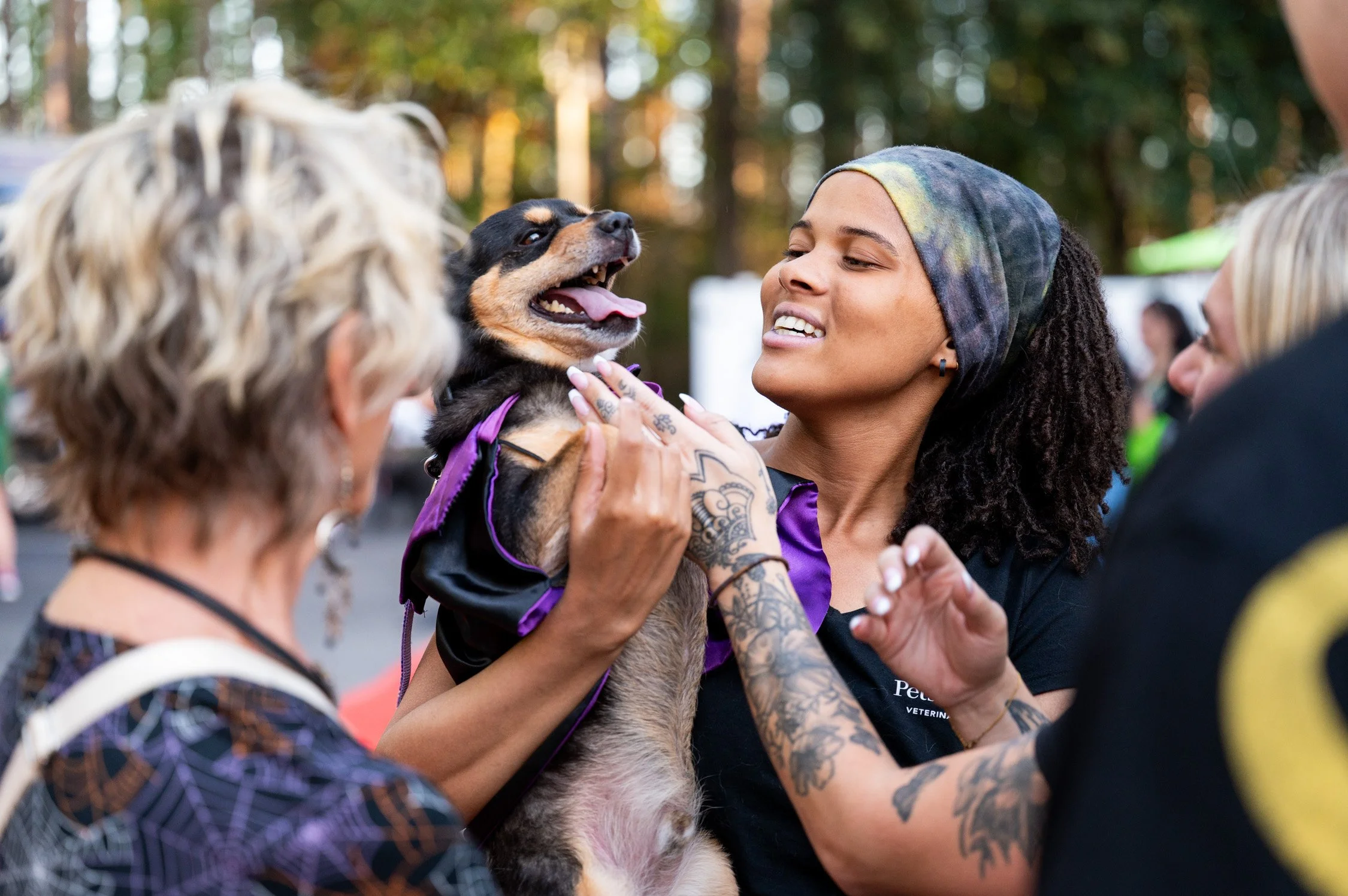 A woman with tattoos and a colorful headscarf holds a happy, black and tan puppy while surrounded by people at an outdoor event.