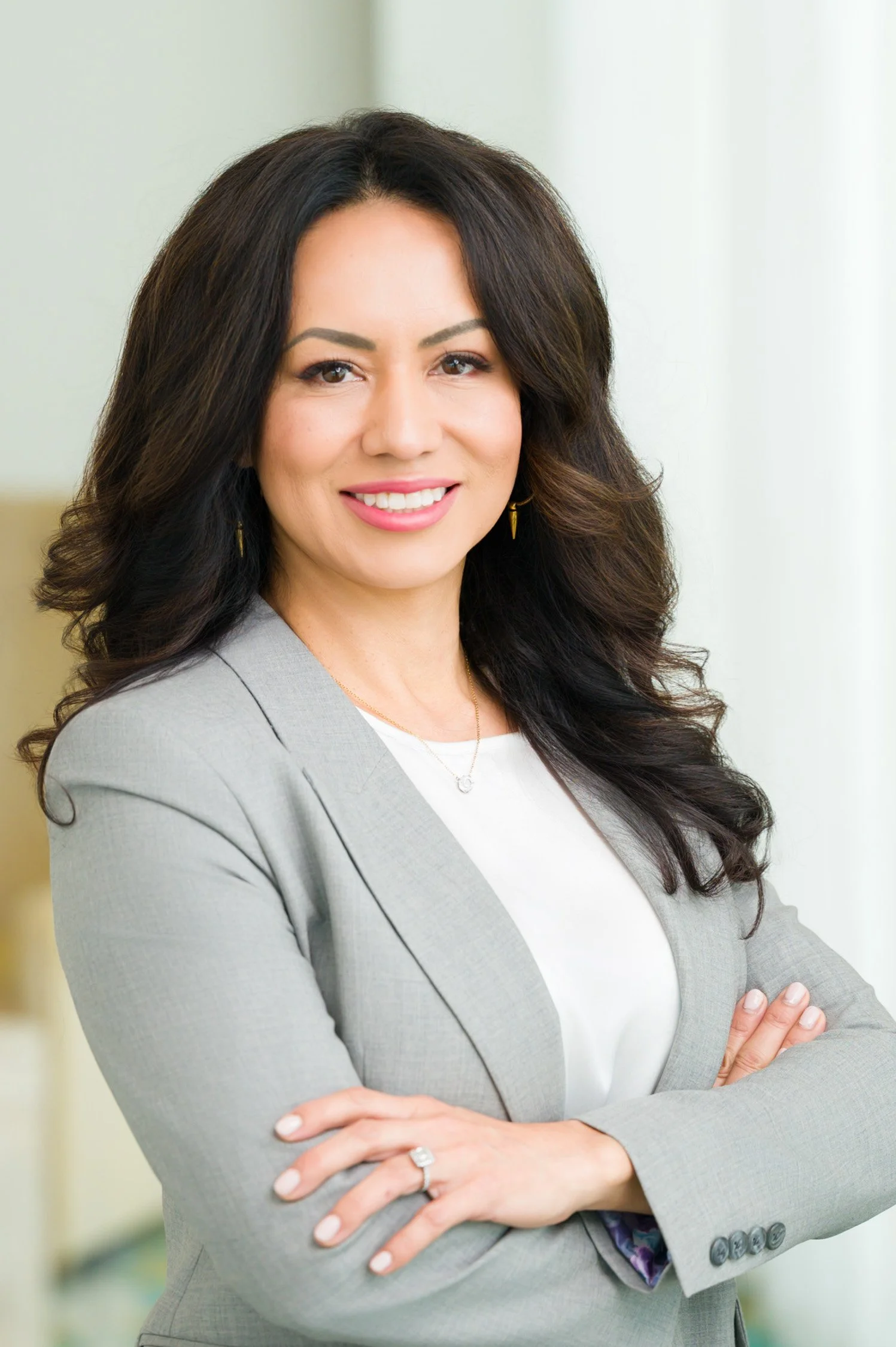 Portrait of a woman with dark, wavy hair, wearing a light gray blazer and white top, standing with arms crossed and smiling at the camera in a professional setting.