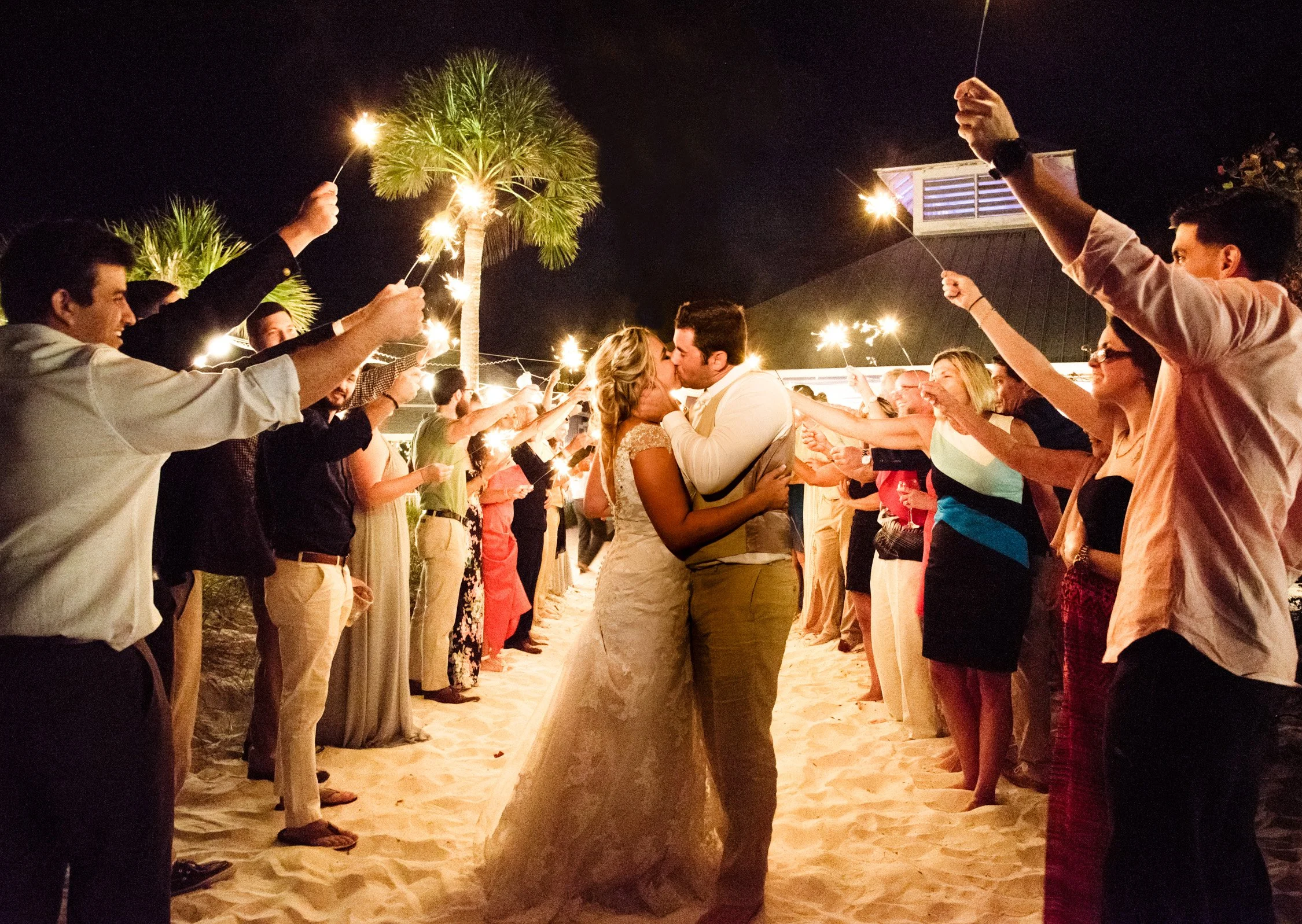 A couple in wedding attire kissing at night on a sandy beach, surrounded by people holding sparklers in celebration.