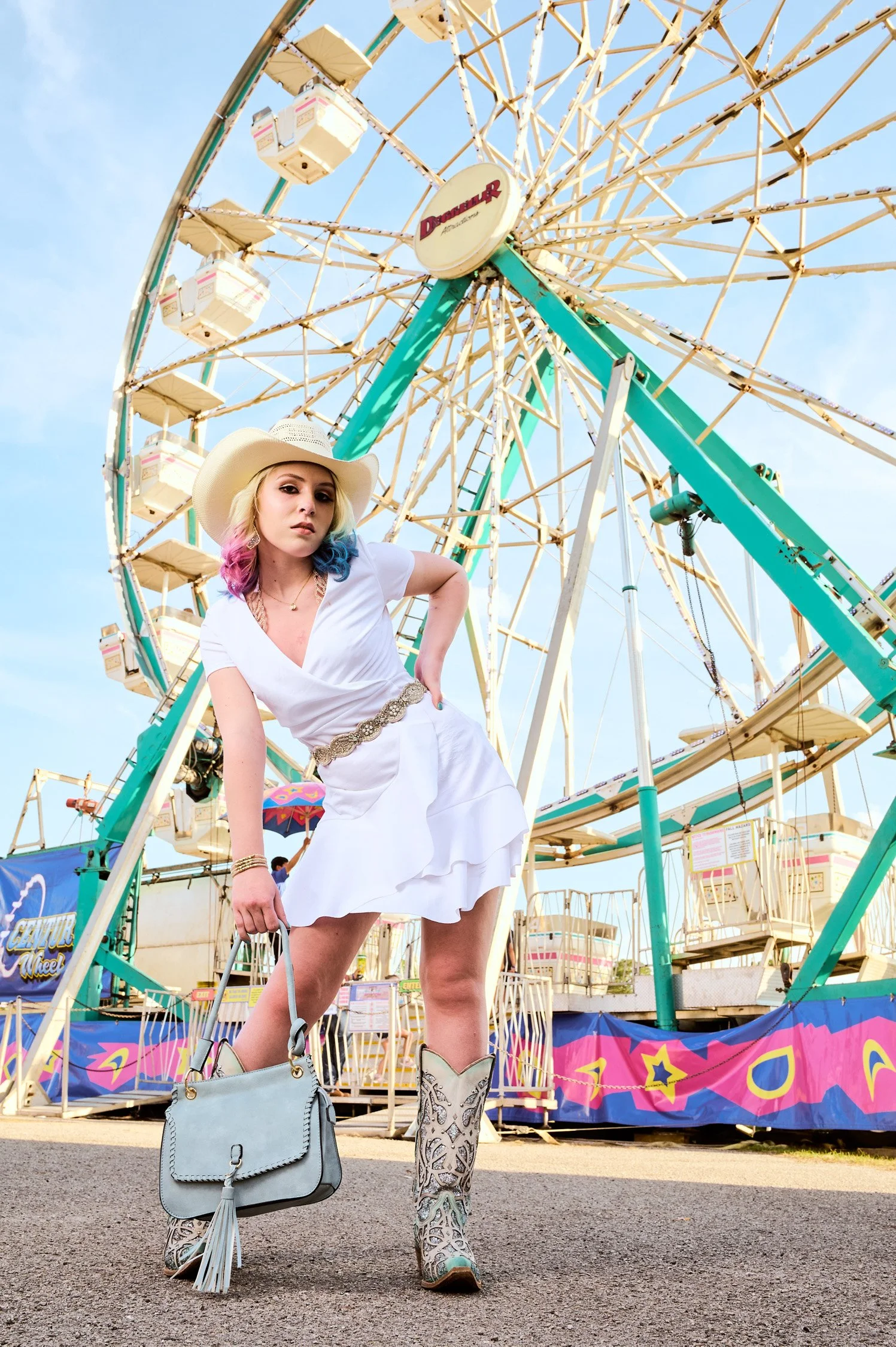 A young woman with blonde hair with pink and blue tips wearing a white dress, a wide-brimmed hat, and cowboy boots standing in front of a Ferris wheel at an amusement park.