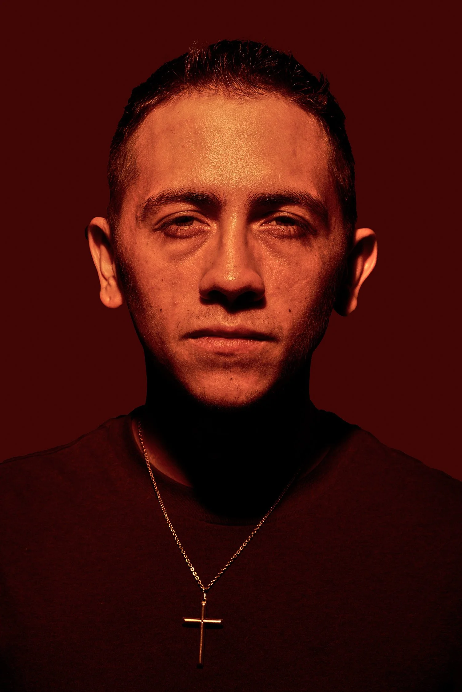 A close-up portrait of a young man with short dark hair, wearing a necklace with a cross pendant, against a dark red background.