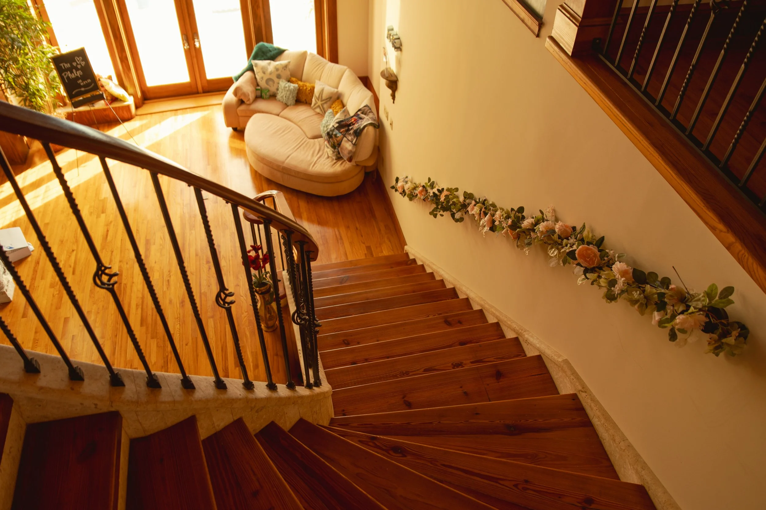 View of a staircase overlooking a living room with wooden flooring, a curved cream sofa with pillows, a chalkboard sign, and an open door with sunlight coming through, decorated with a flower garland along the wall.