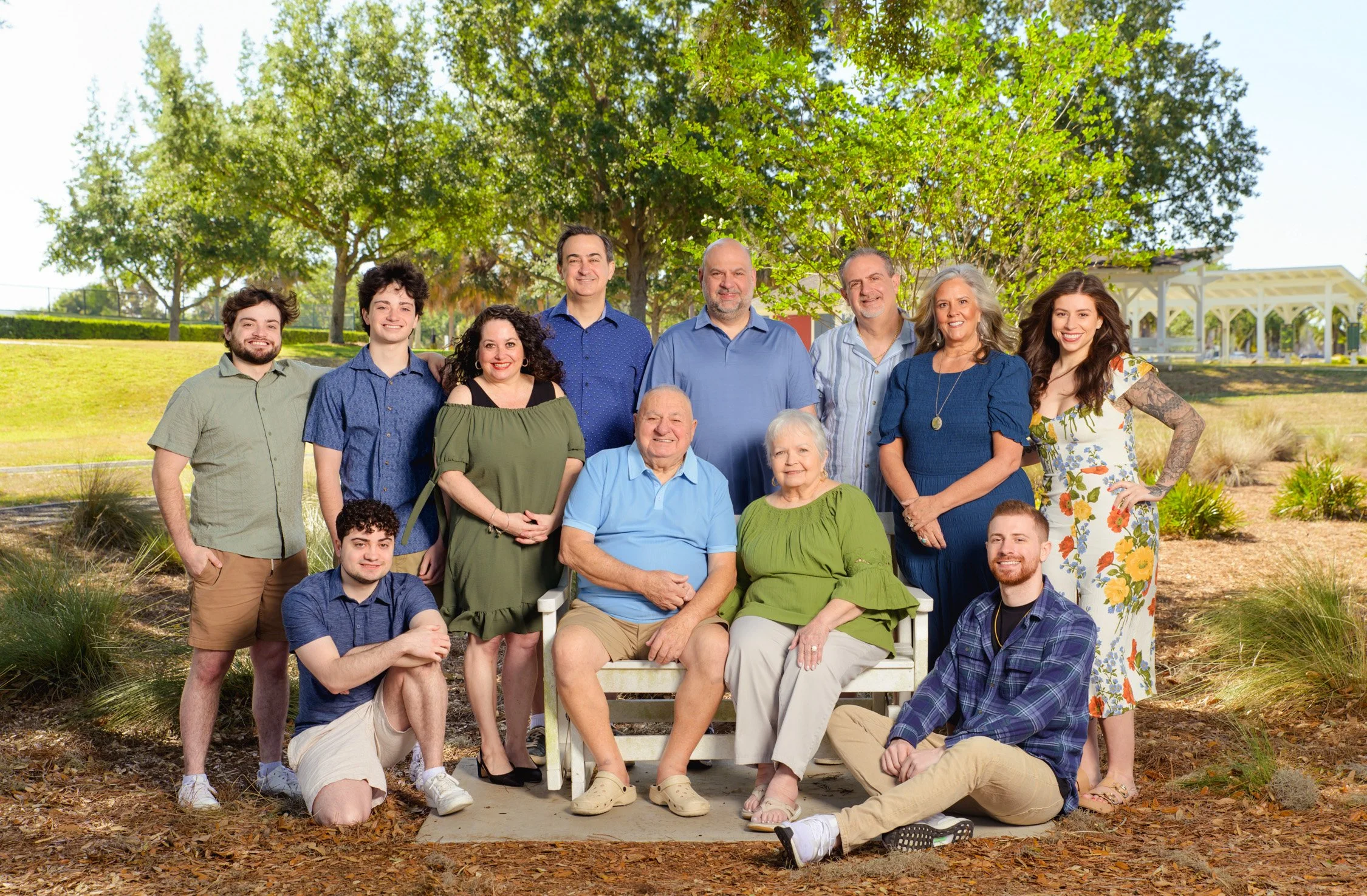 A large extended family gathered outdoors for a group photo, with trees and a pavilion in the background.