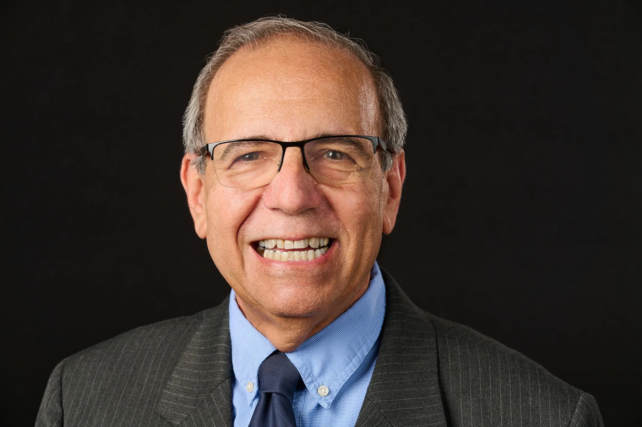 A middle-aged man smiling, wearing glasses, a dark pinstripe suit, a light blue shirt, and a dark tie, against a black background.