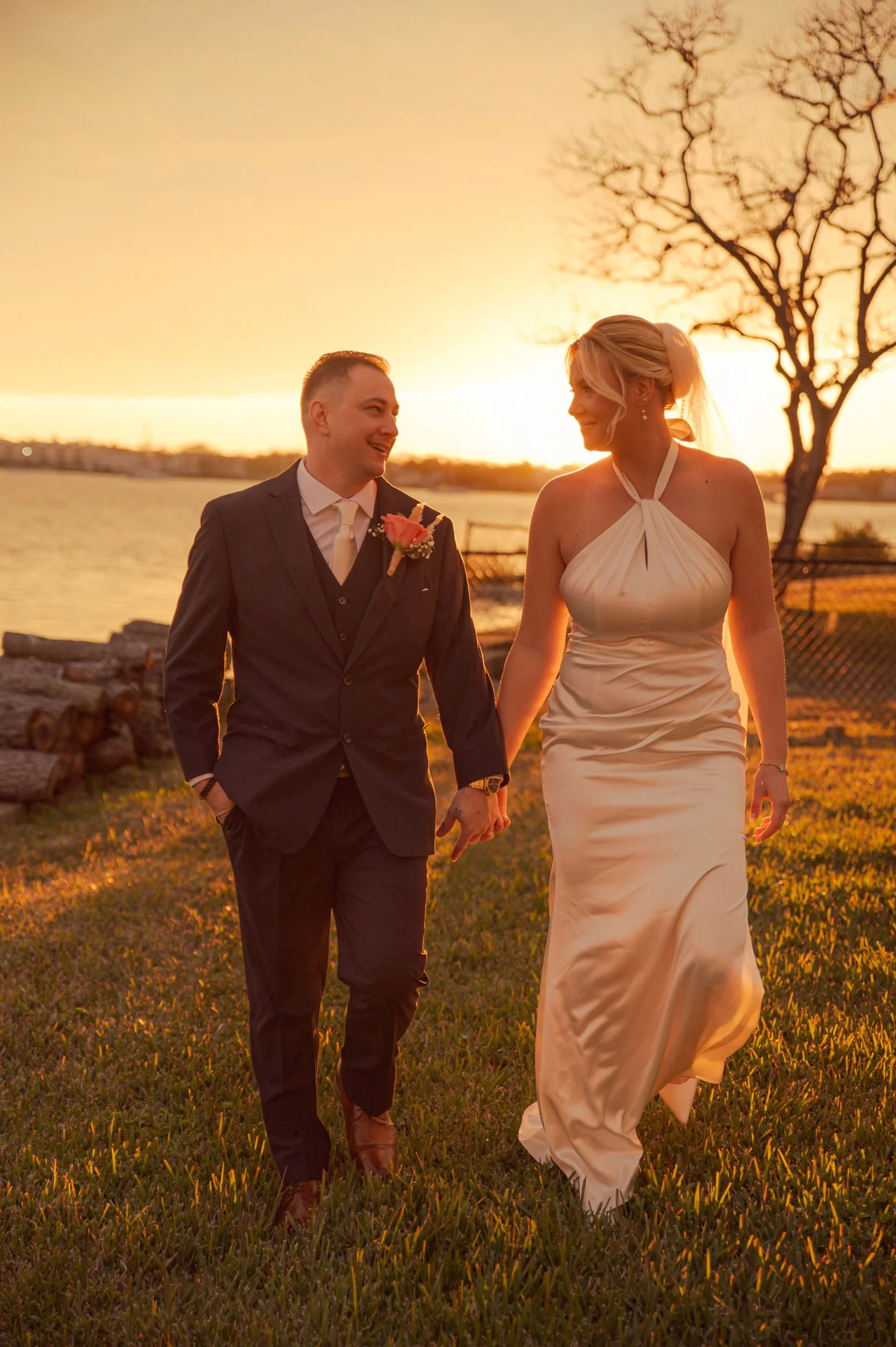 A newlywed couple holding hands and walking outdoors during sunset, with a body of water, logs, and a leafless tree in the background.