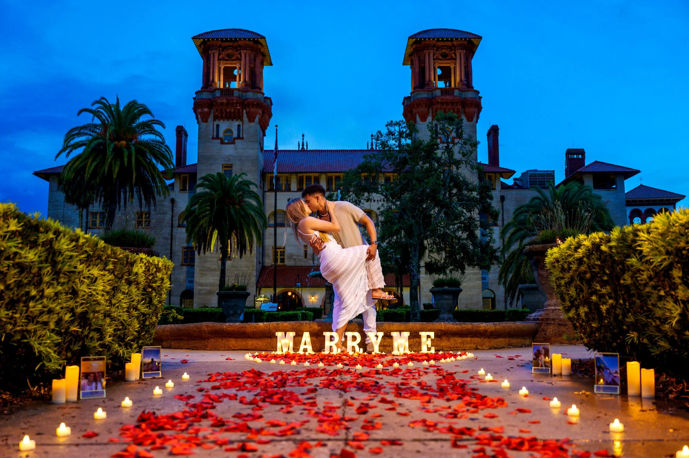 A romantic couple kissing in front of a historic castle during twilight, with candles, rose petals, and illuminated letters spelling "MARRY ME" on the ground.