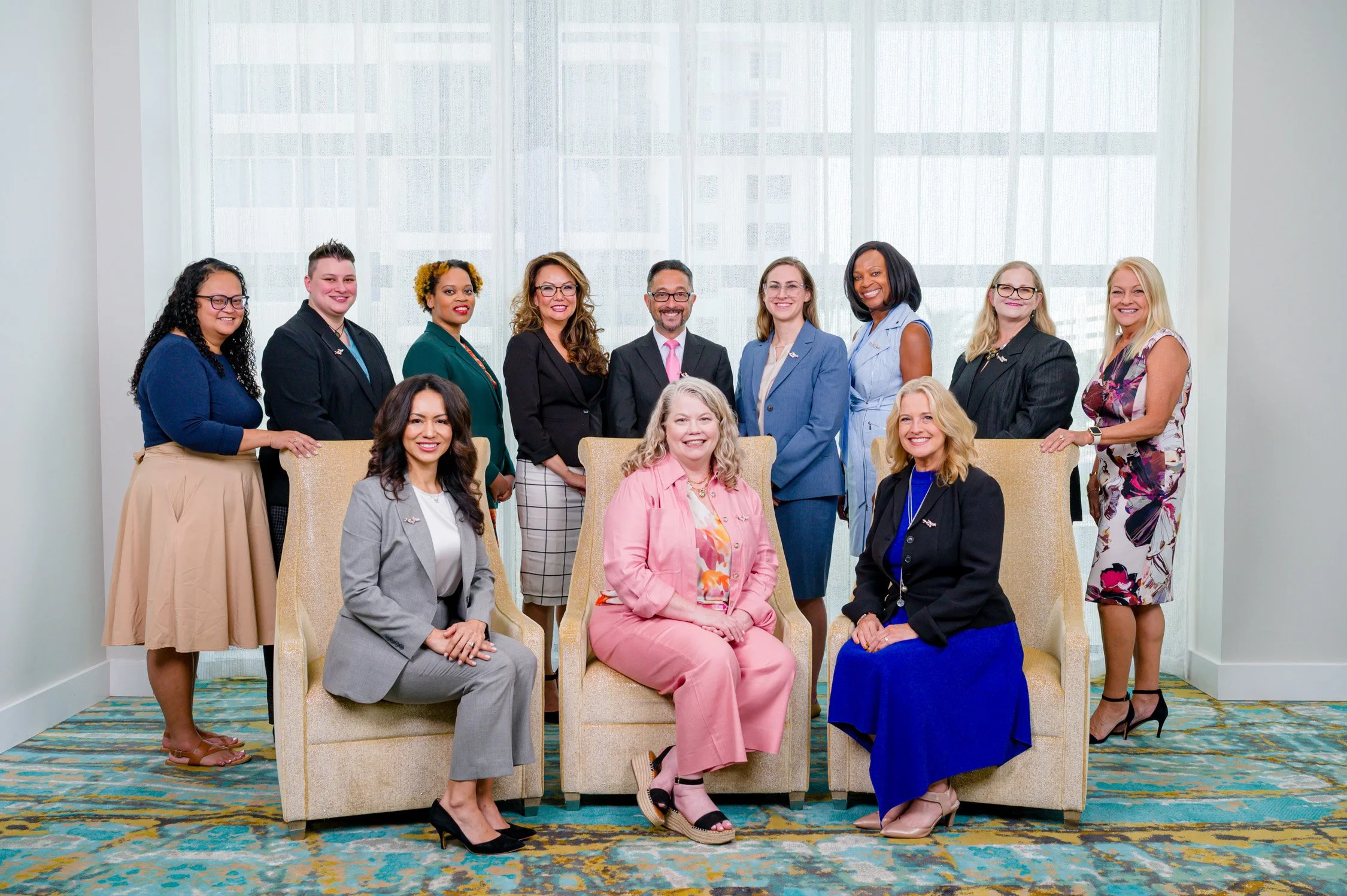 A diverse group of women and one man in professional attire posing in a bright office space with large windows, some sitting and some standing.