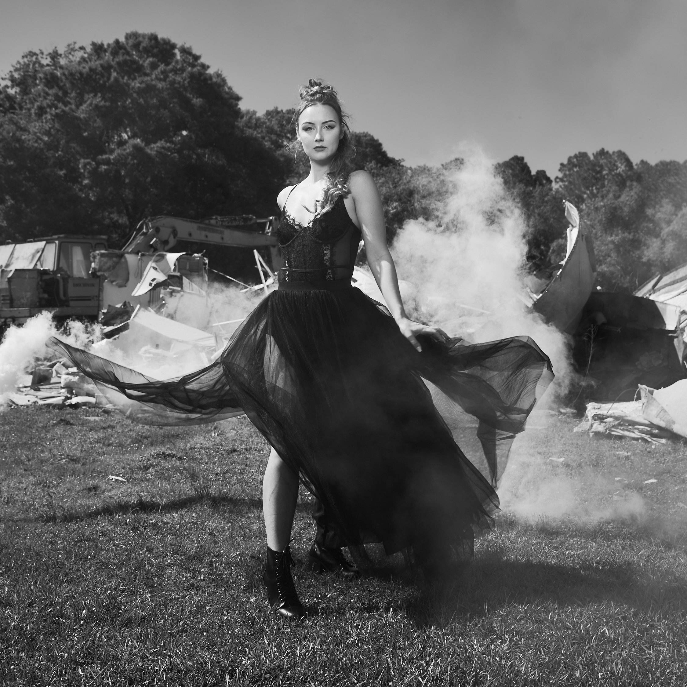 A woman in a black dress stands in a grassy field with wreckage and smoke behind her.