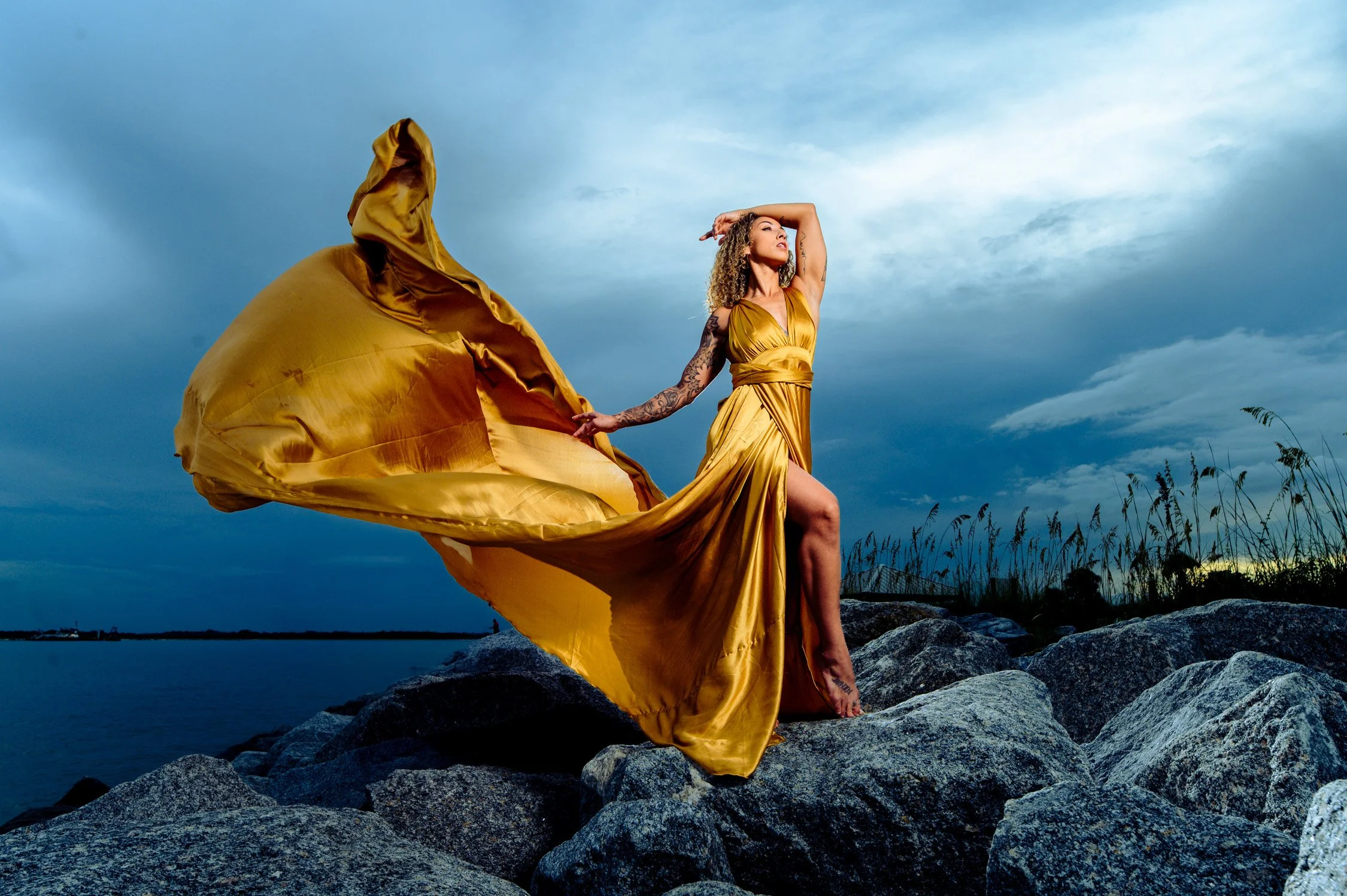 Woman in a flowing gold dress standing on rocks by water with a cloudy sky in background.