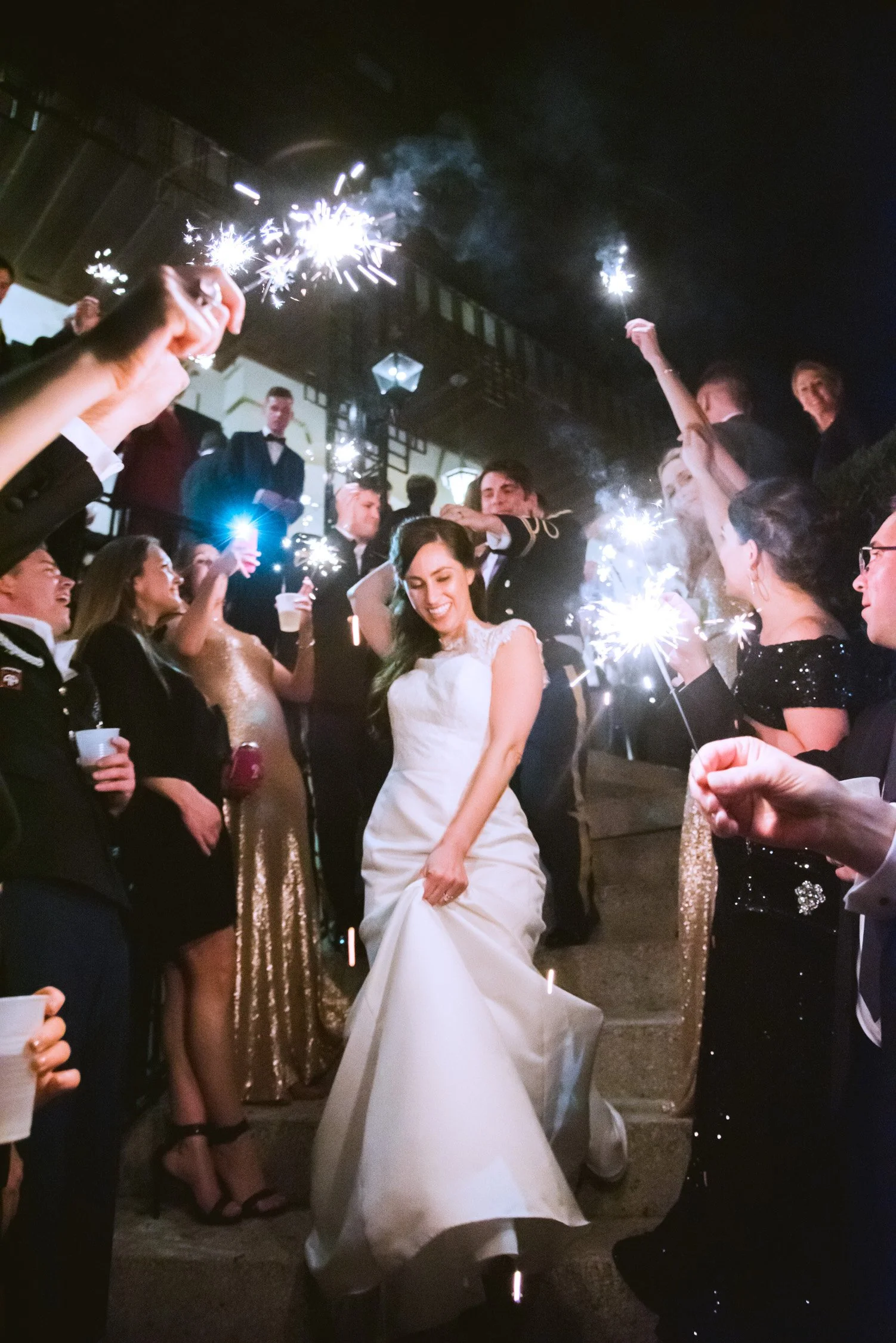 A bride in a white wedding gown standing on stairs surrounded by guests holding sparklers, celebrating at a wedding reception.