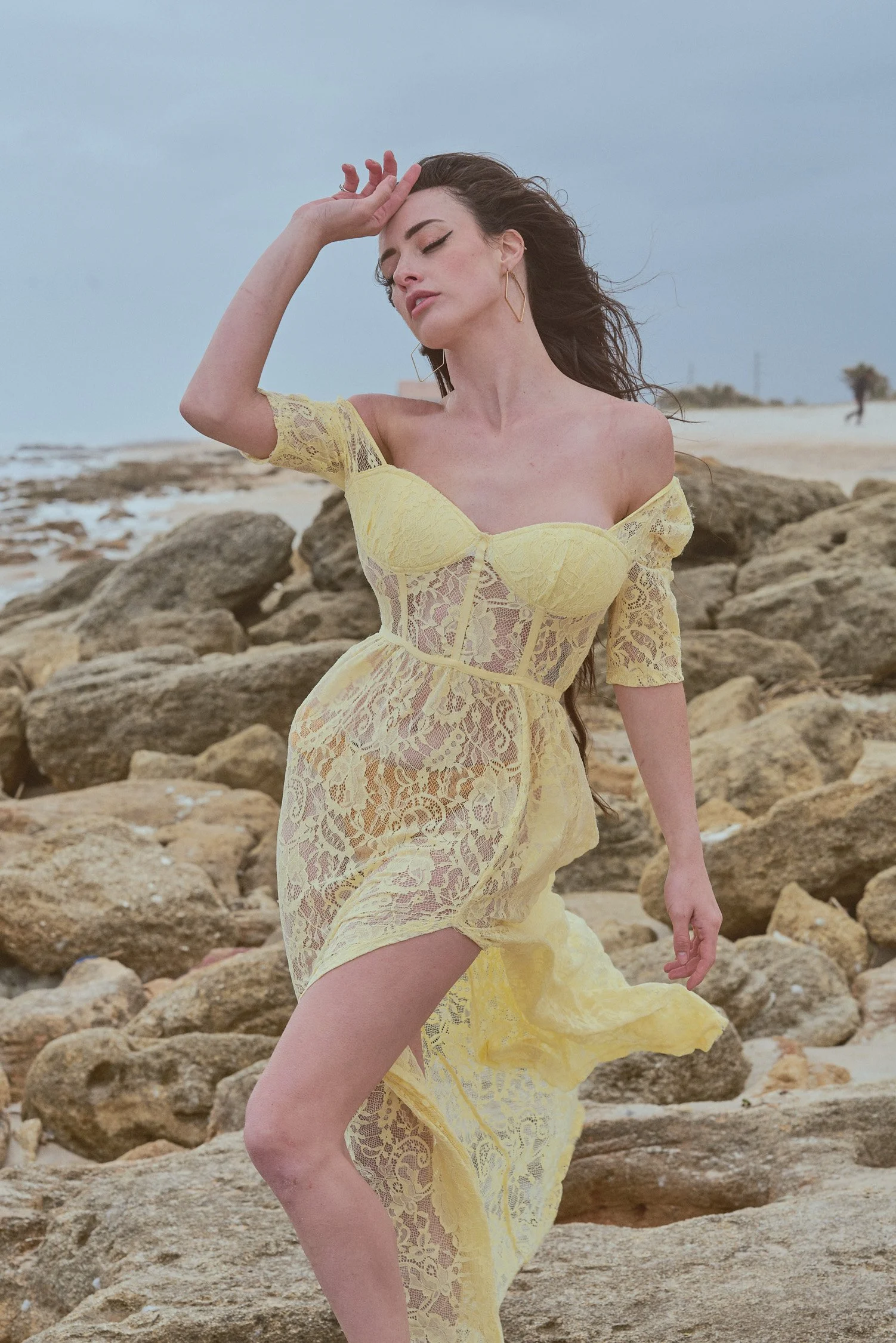 A woman in a yellow lace dress standing on a rocky beach, with her eyes closed and one hand on her forehead.