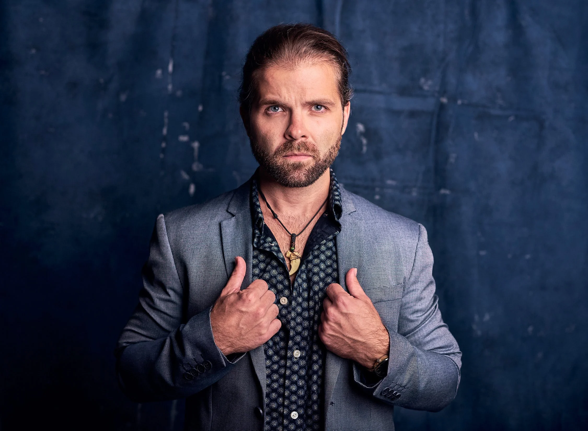 A man with styled hair and a beard wearing a gray blazer over a patterned shirt, standing against a dark textured background, looking directly at the camera with his hands adjusting his blazer.