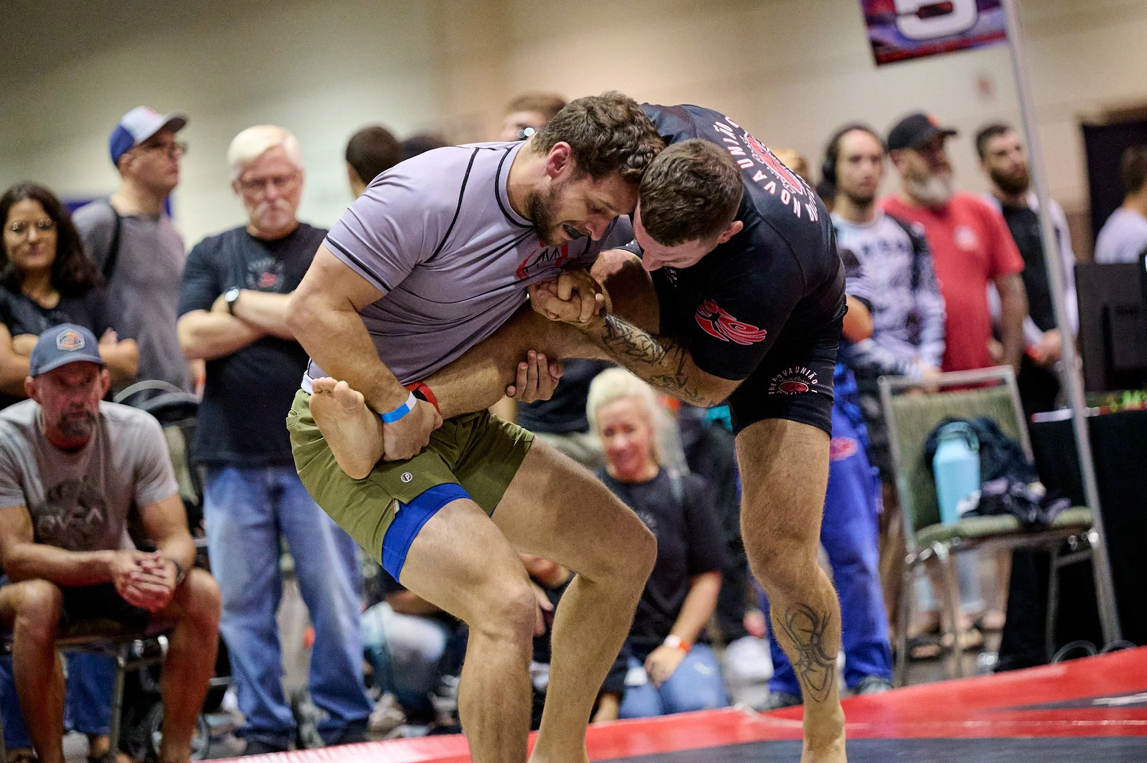 Two men engaged in a grappling match at a wrestling event, with spectators watching around them.