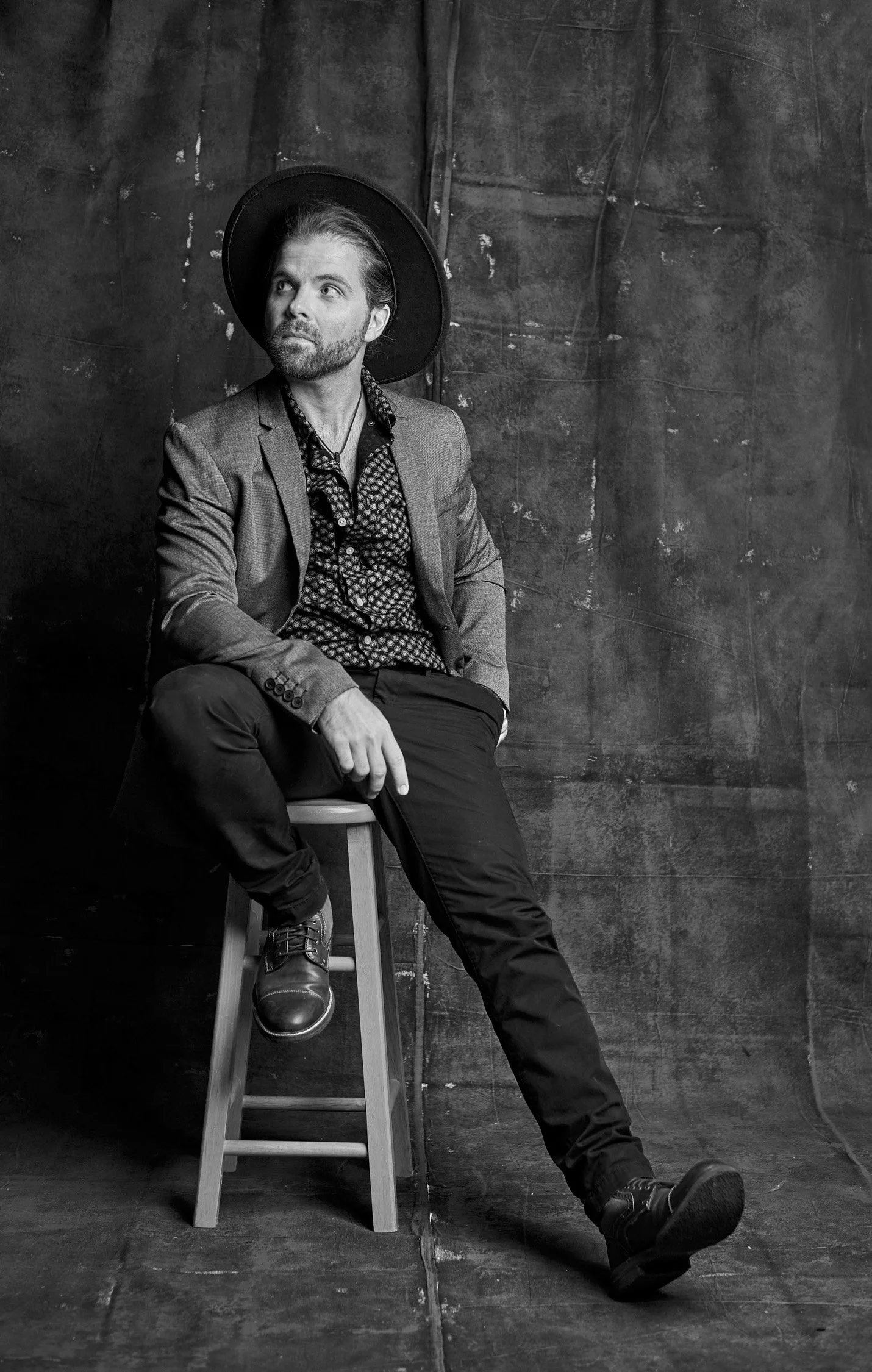 A man sitting on a wooden stool, wearing a blazer, patterned shirt, czapka, and boots, looking to the side in a studio with a textured dark background, in black and white.