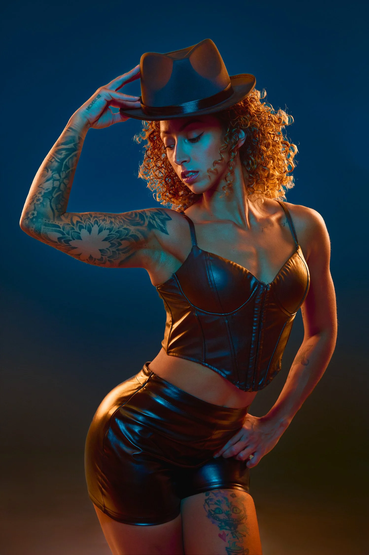 A woman with curly red hair, wearing a black leather cropped top and matching leather shorts, poses with a fedora hat, her hand on her hip, against a gradient background.
