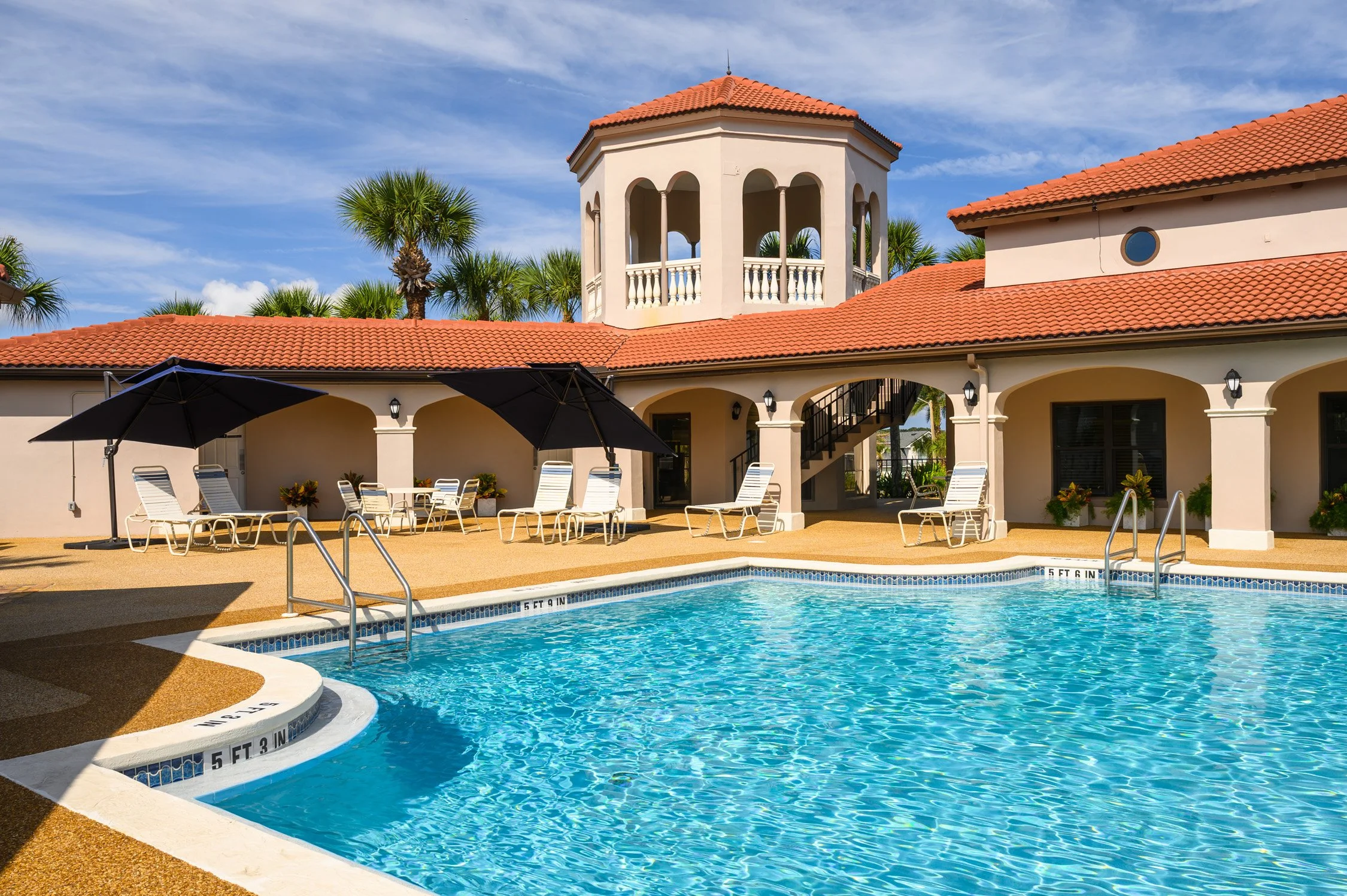 Luxury resort pool area with lounge chairs, umbrellas, and a building with arches and red-tiled roof, surrounded by palm trees under a blue sky.
