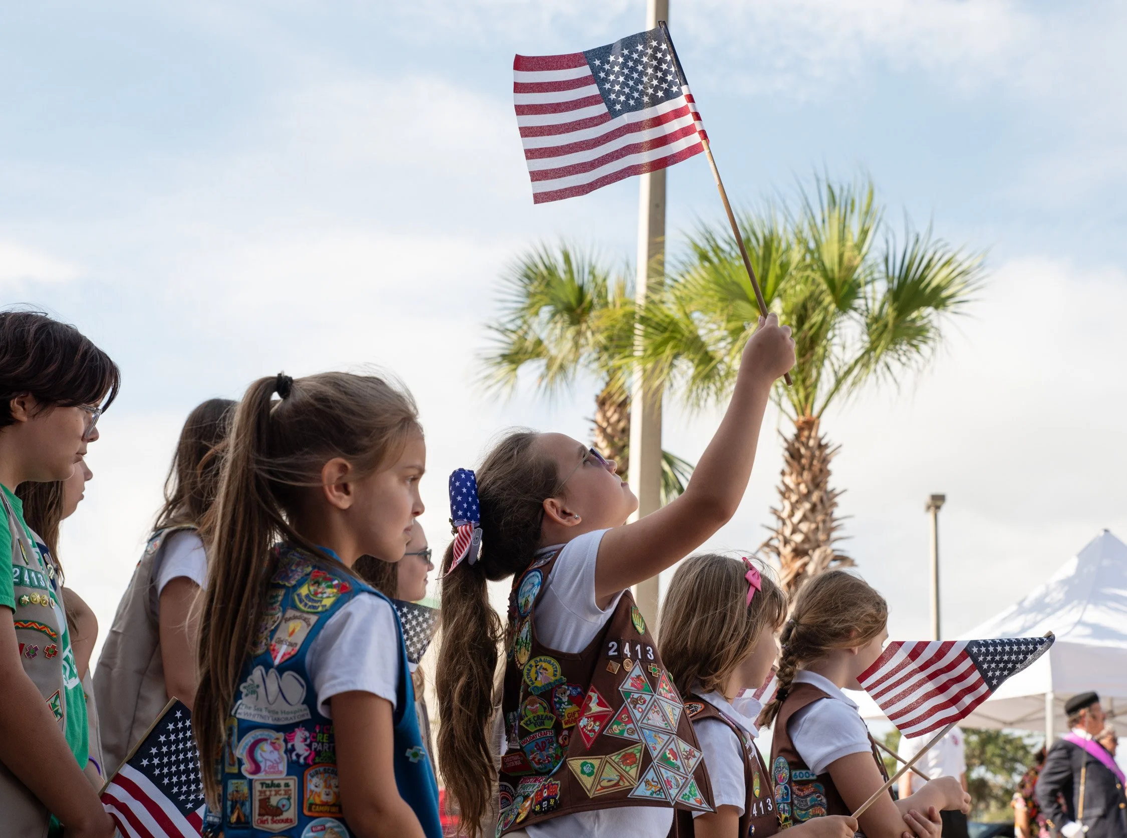 Girls in Girl Scout uniforms holding American flags at an outdoor event with palm trees in the background.
