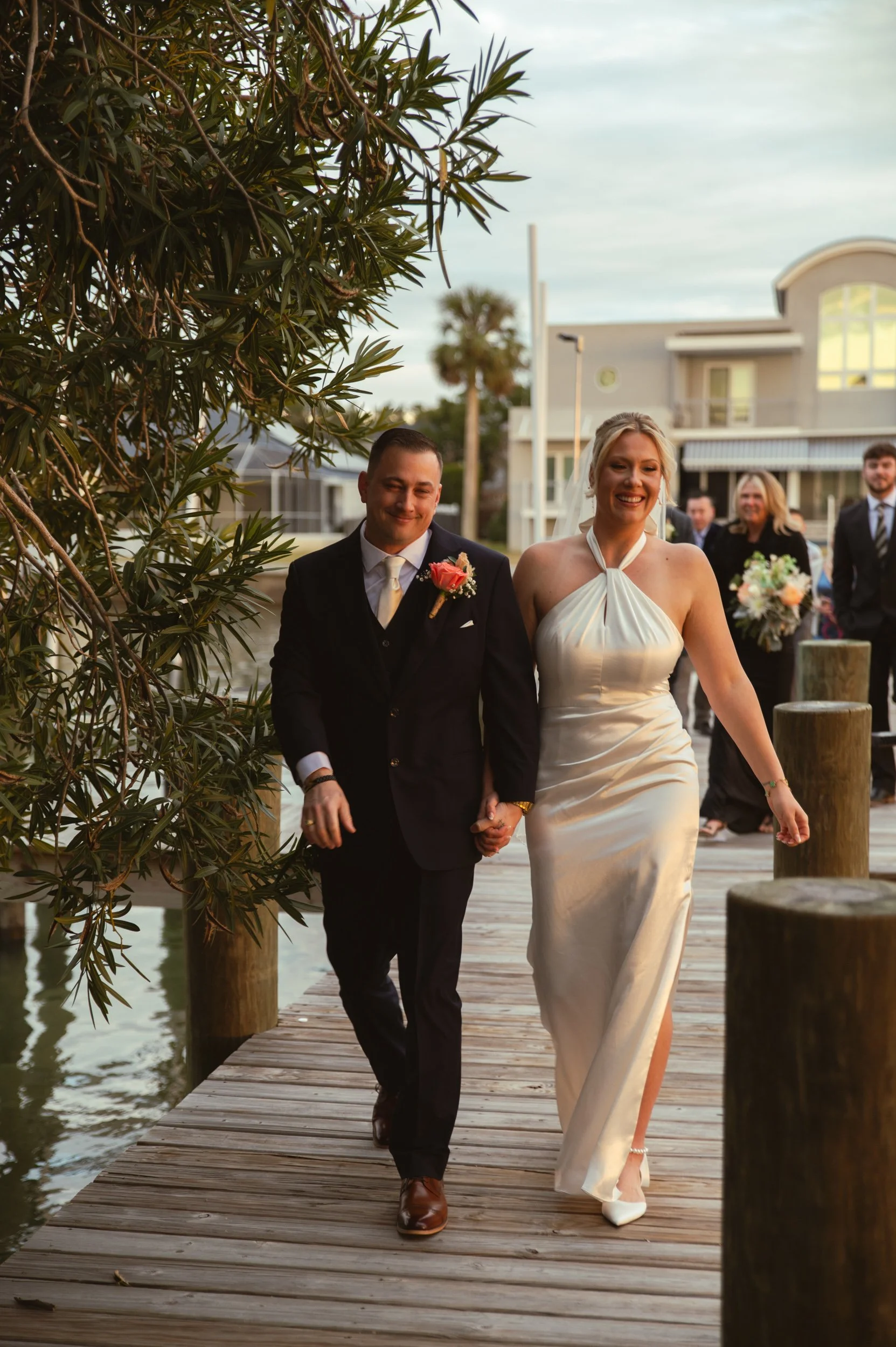 A newlywed couple walking hand in hand on a wooden dock, smiling. The groom is in a black suit with a pink boutonniere, and the bride is in a cream-colored halter wedding gown. In the background, friends and family are gathered, with a white building
