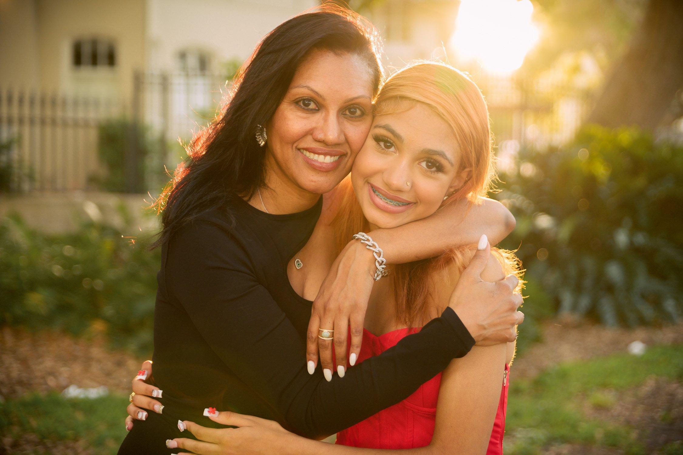 Two women embracing outdoors at sunset, smiling, with a house and trees in the background.