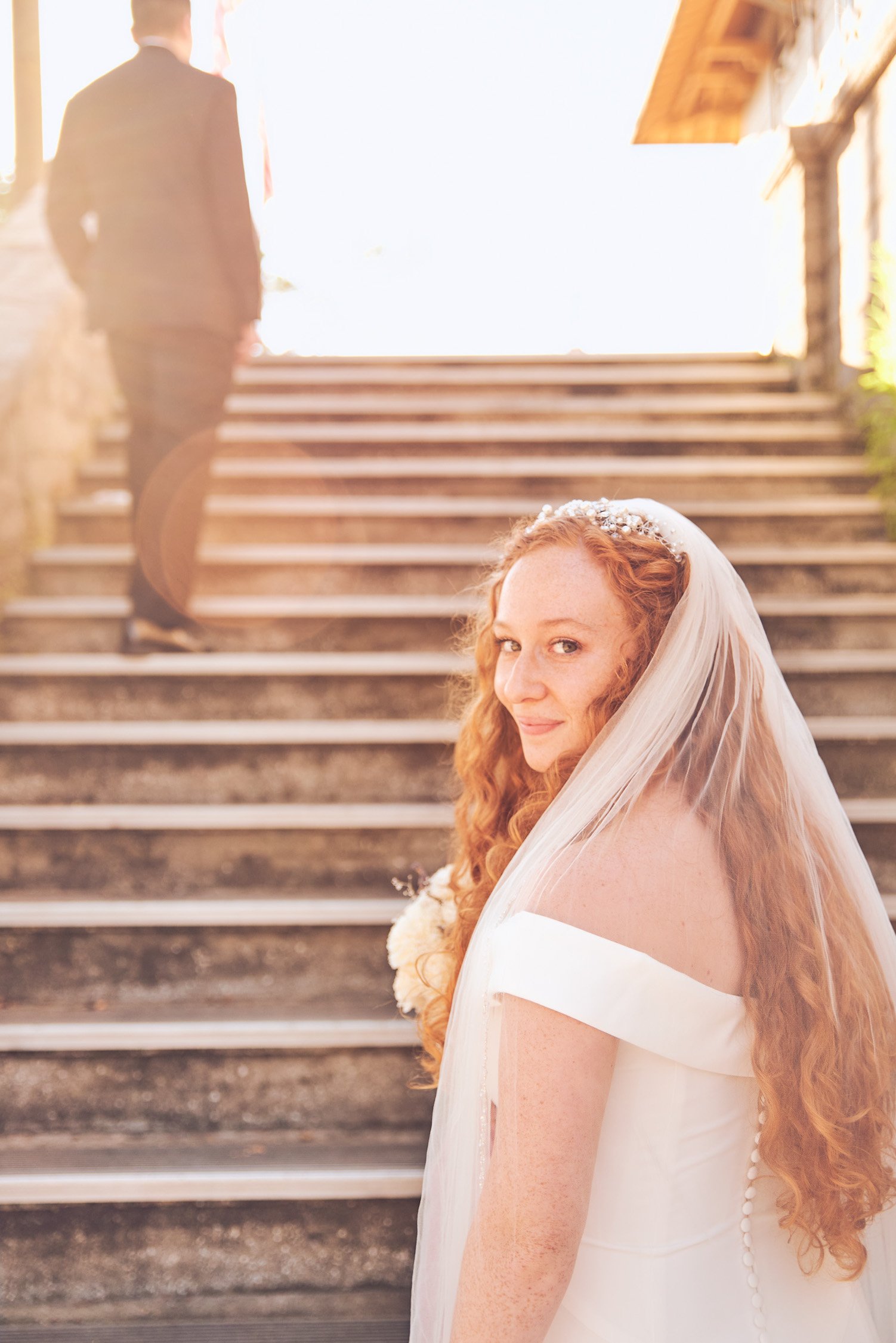 A bride with red curly hair, wearing a white dress and veil, standing on outdoor stairs during sunset, looking over her shoulder with a smile, with a man in a dark suit walking away in the background.