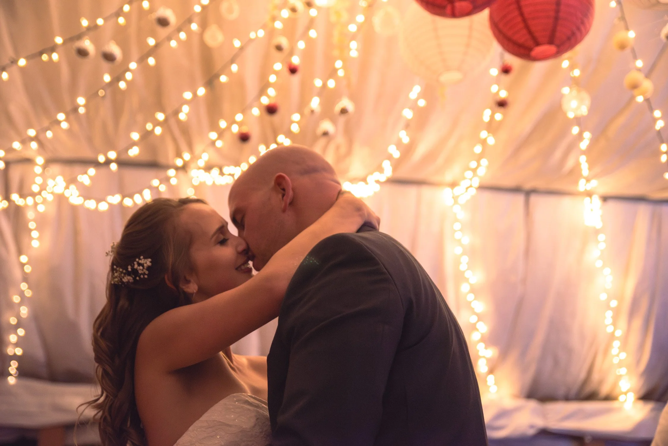 A bride and groom kiss under warm string lights and pink paper lanterns in a decorated tent.