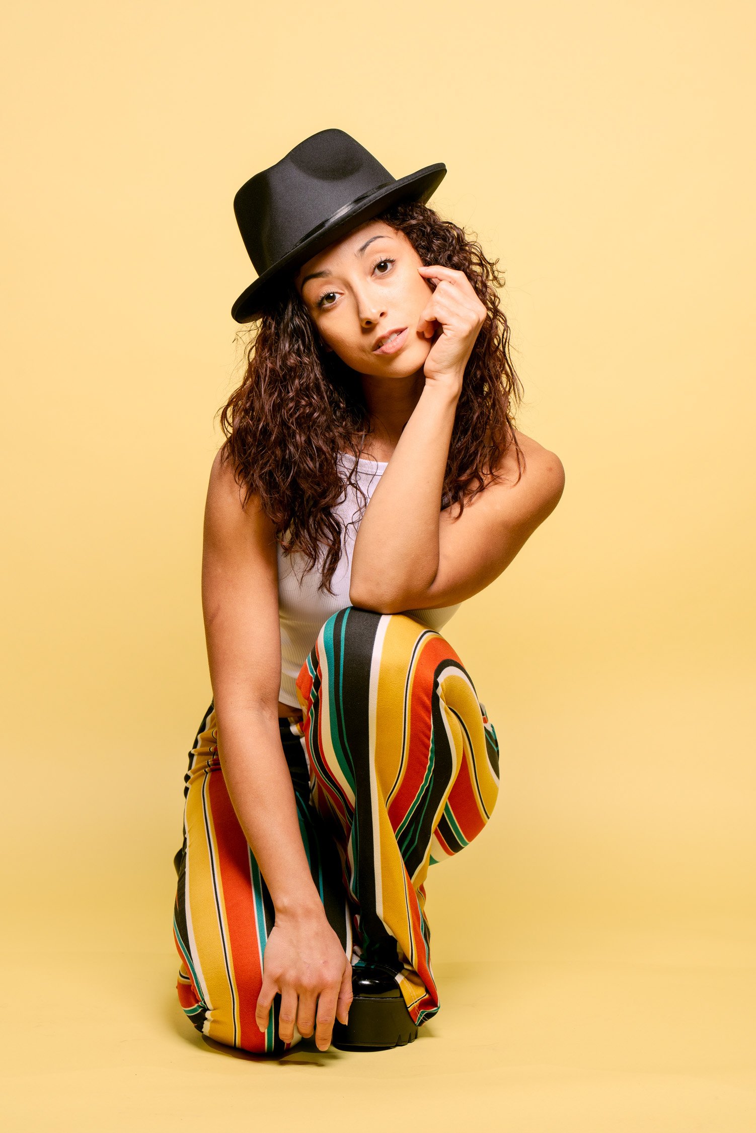 A young woman with curly hair wearing a black hat and striped colorful pants, posing against a yellow background.