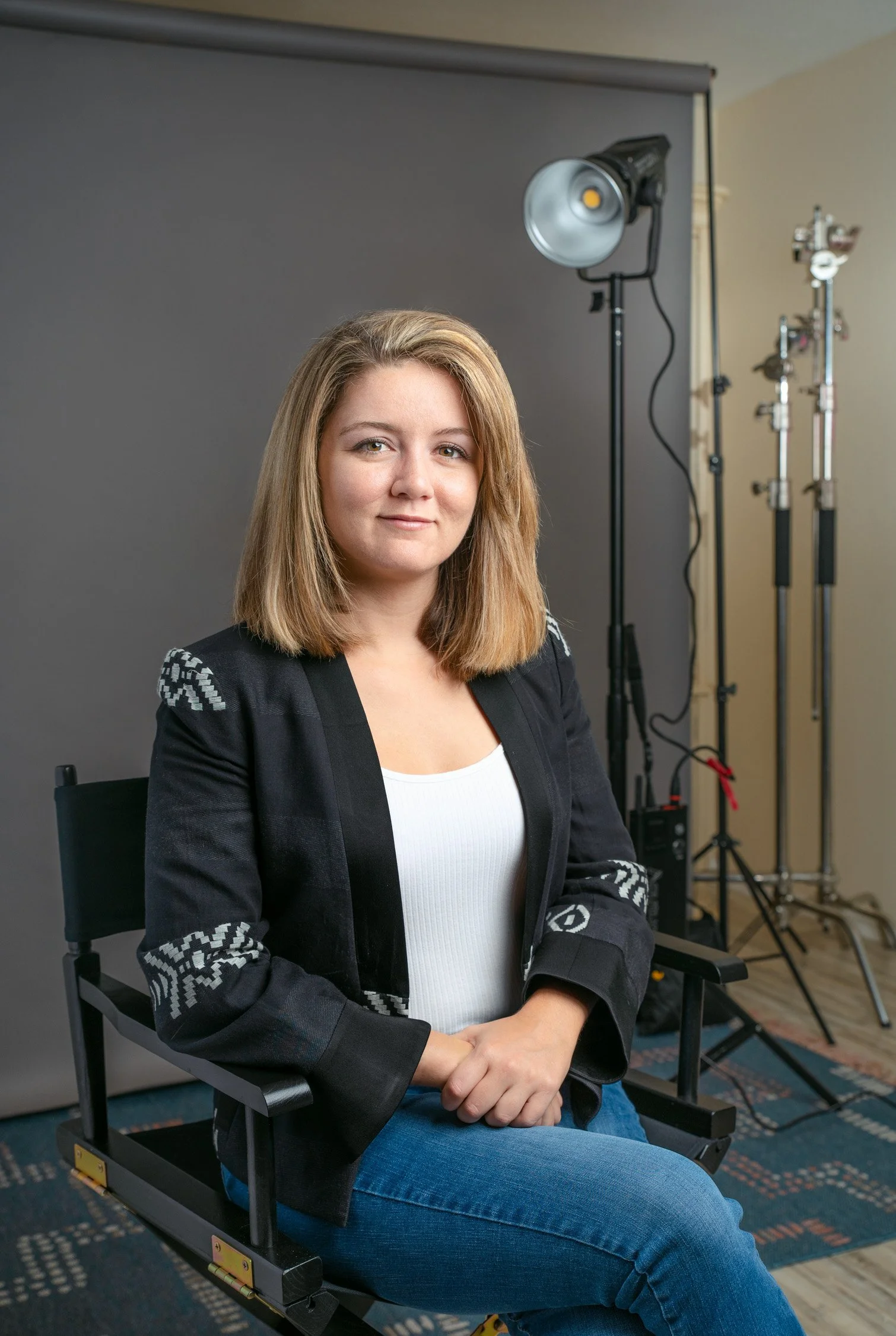 A woman with shoulder-length blonde hair sitting on a director's chair in a photography studio with professional lighting equipment and a gray backdrop.