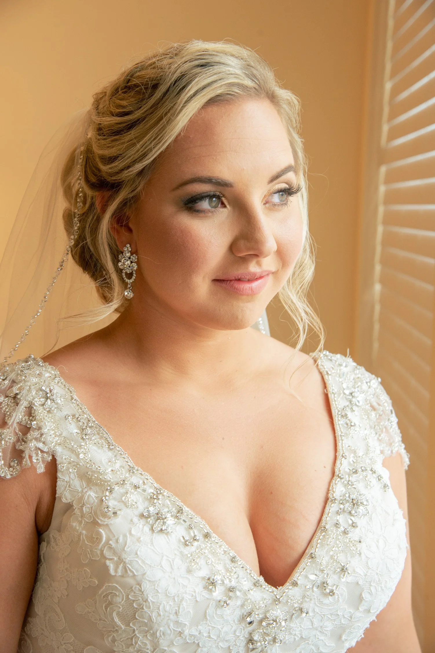 Close-up of a bride looking to the side, wearing a wedding dress with intricate beading detail, earrings, and a veil, standing near window blinds.