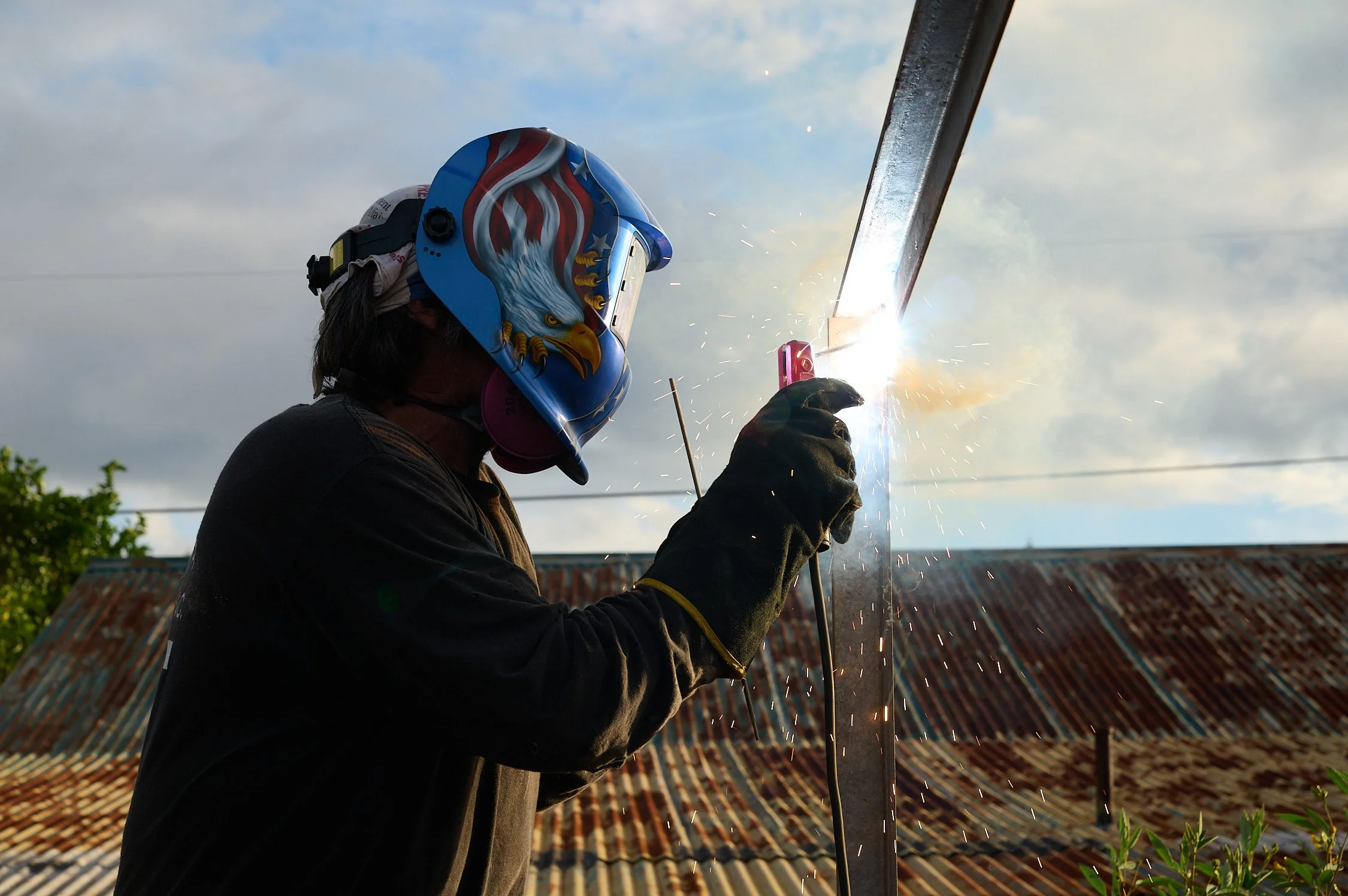 A person welding metal outdoors, wearing a colorful helmet with an eagle design, gloves, and protective gear, sparks flying from the welding process, with a rusted roof and cloudy sky in the background.
