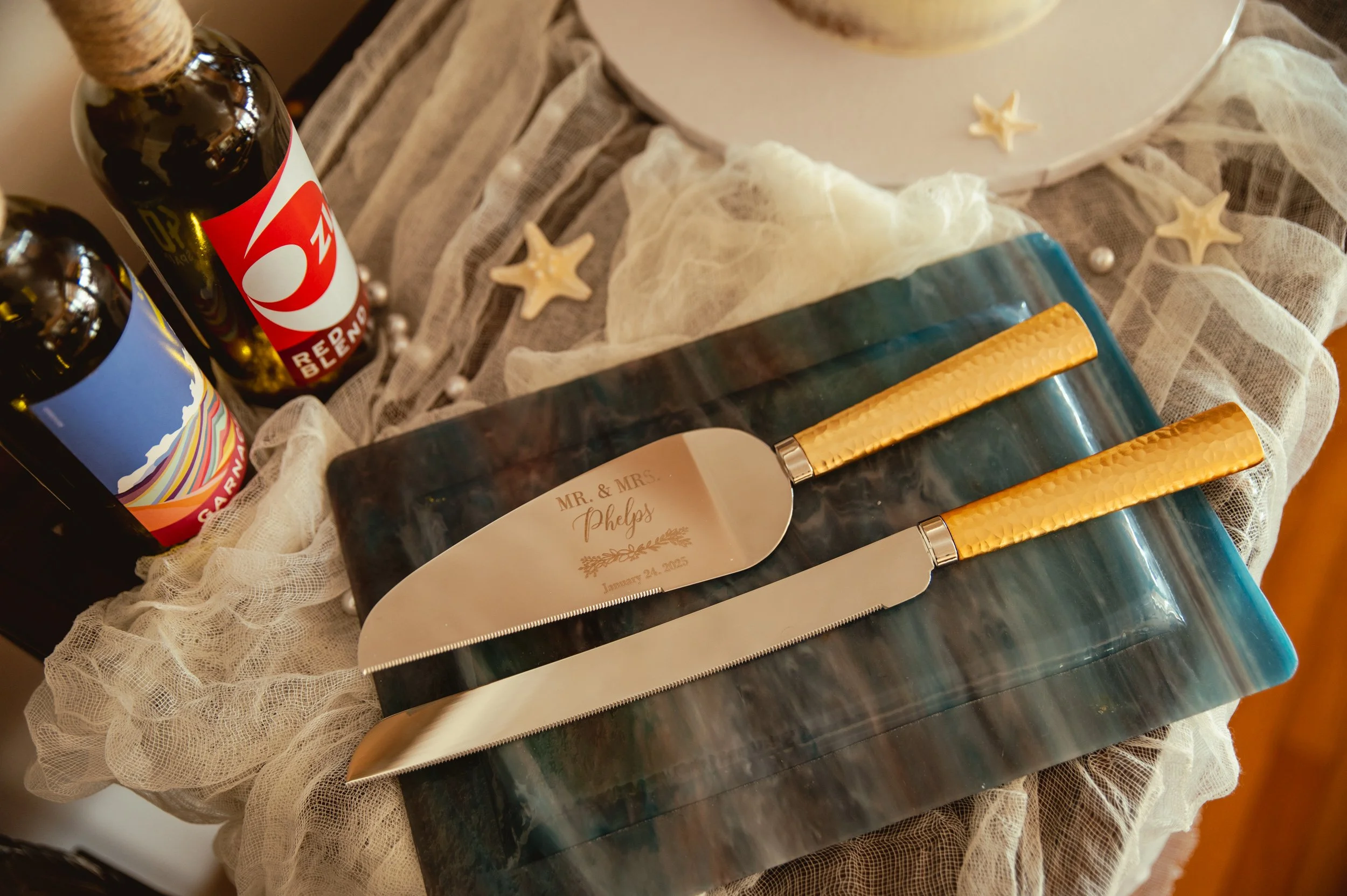 Decorative wedding cake serving set with engraved names and date, bottles of wine or champagne, and starfish decorations on a table with lace and fabric.