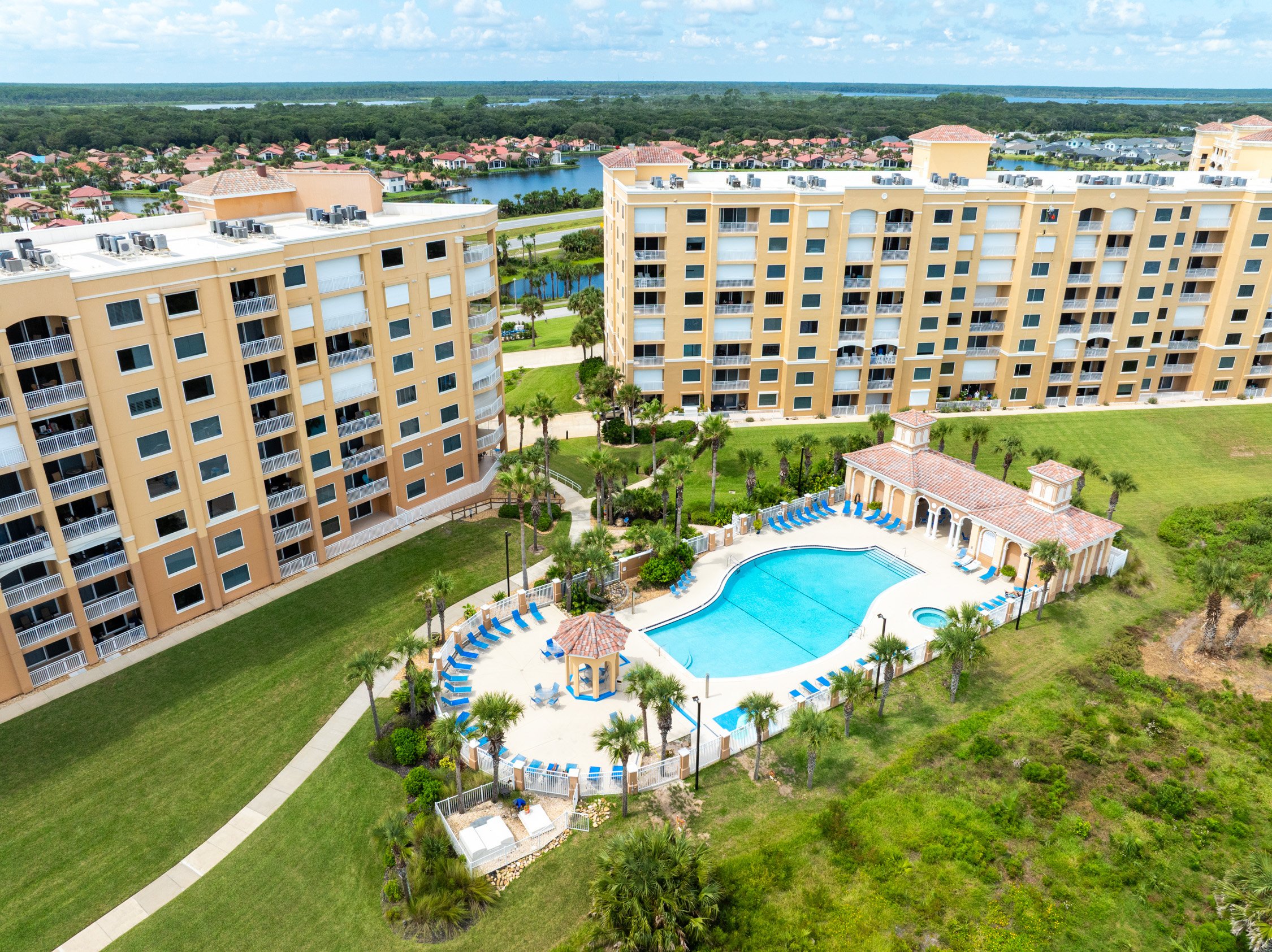 Aerial view of a resort-style apartment complex with yellow buildings, a large swimming pool, poolside lounge chairs, and surrounding greenery, lakes, and additional housing in the background.