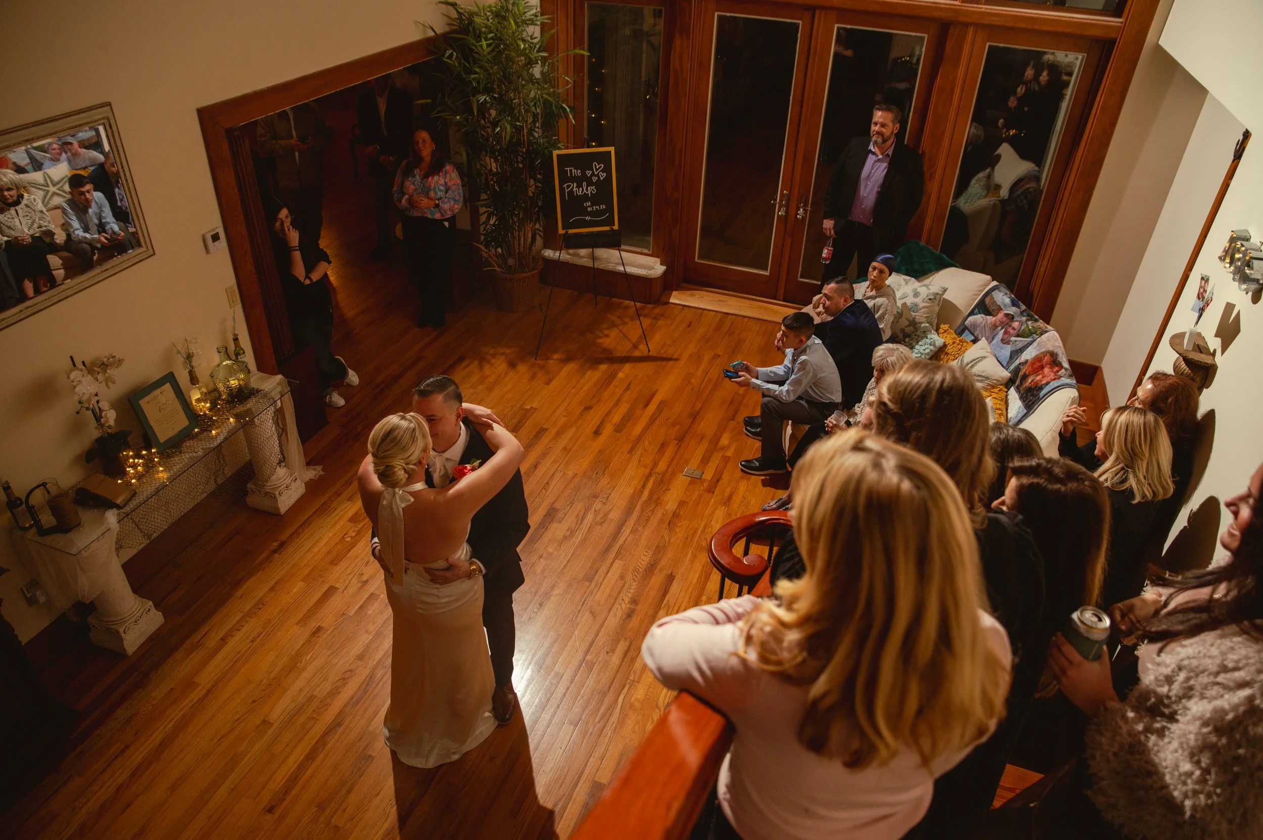 A wedding reception with a bride and groom dancing while guests watch. The room has wooden floors, a large window with brown frames, and a cozy sofa. Guests are seated, some are standing, and one person is playing a video game. Decorations include a 