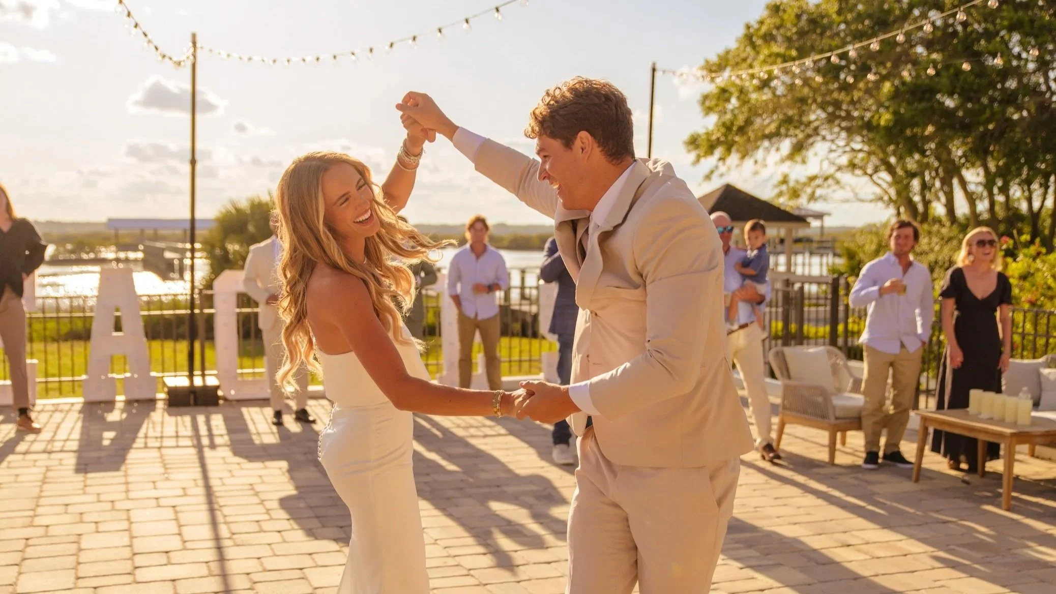 A couple is dancing outdoors at a wedding celebration during sunset, surrounded by guests. The woman has long curly blonde hair and is wearing a white dress, and the man has short curly hair and is wearing a light-colored suit. They are holding hands and smiling, with people in the background, some taking photos, on a patio with string lights overhead.