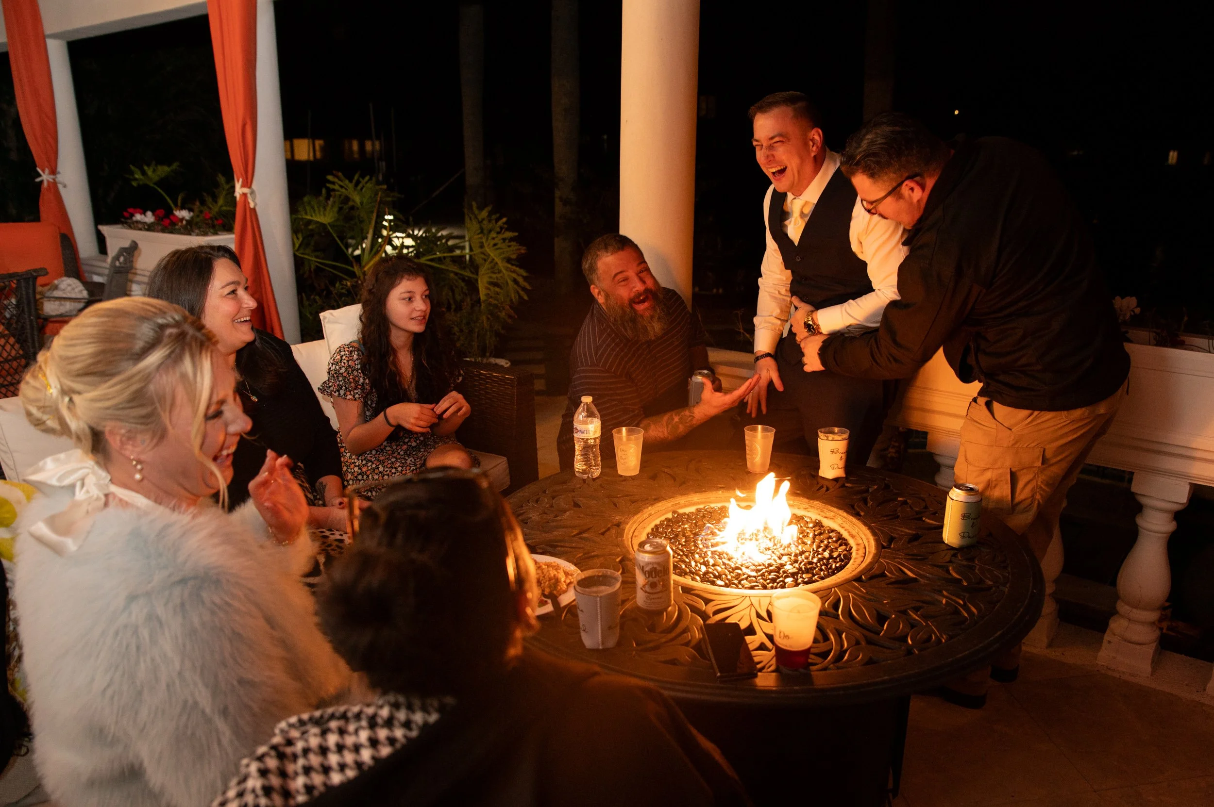 Group of people gathered around a firepit on a porch at night, laughing and enjoying each other's company.