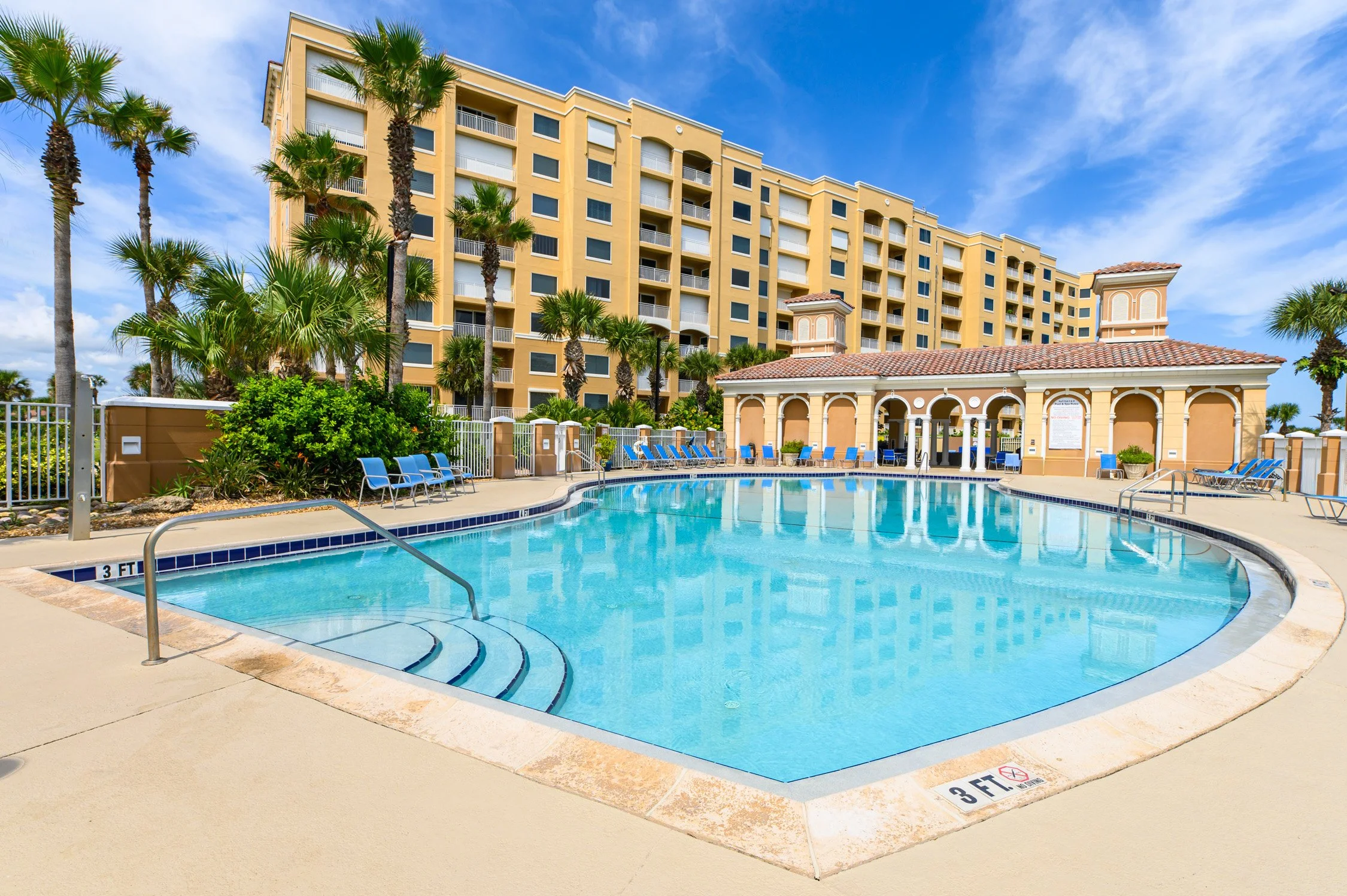 Pool area with blue lounge chairs, surrounding beige concrete, in front of a tall yellow apartment building with balconies, palm trees, and a partly cloudy sky.