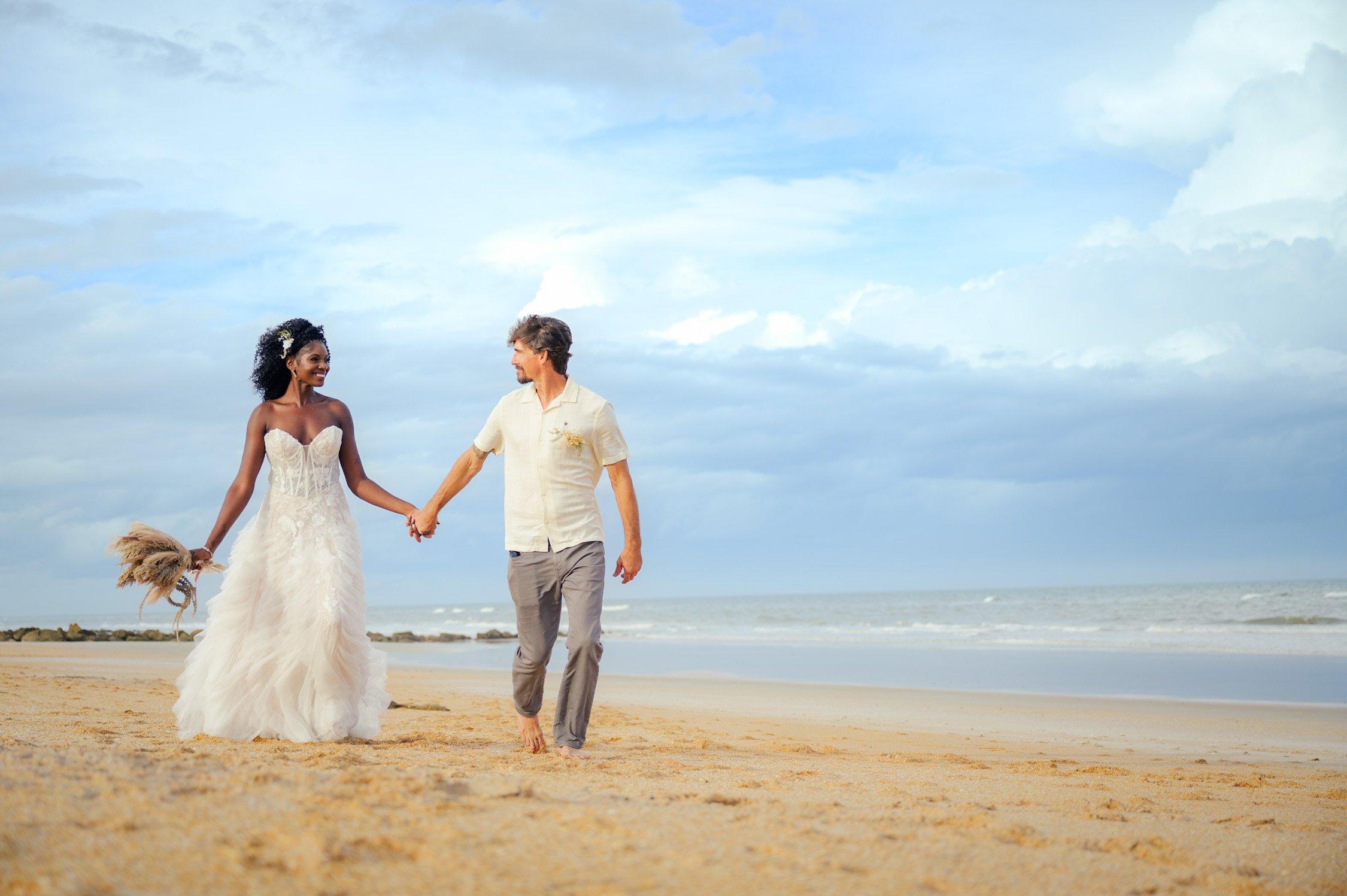 A couple walking on a sandy beach with the ocean in the background, the woman in a white wedding dress holding a bouquet and the man in casual attire.