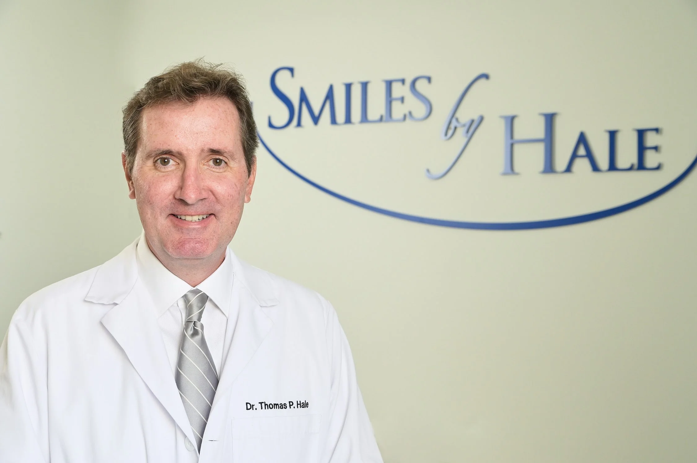 A smiling man in a white coat with a name tag that reads Dr. Thomas P. Hale, standing in front of a light-colored wall with a sign that says "Smiles by Hale" and a curved line underneath.