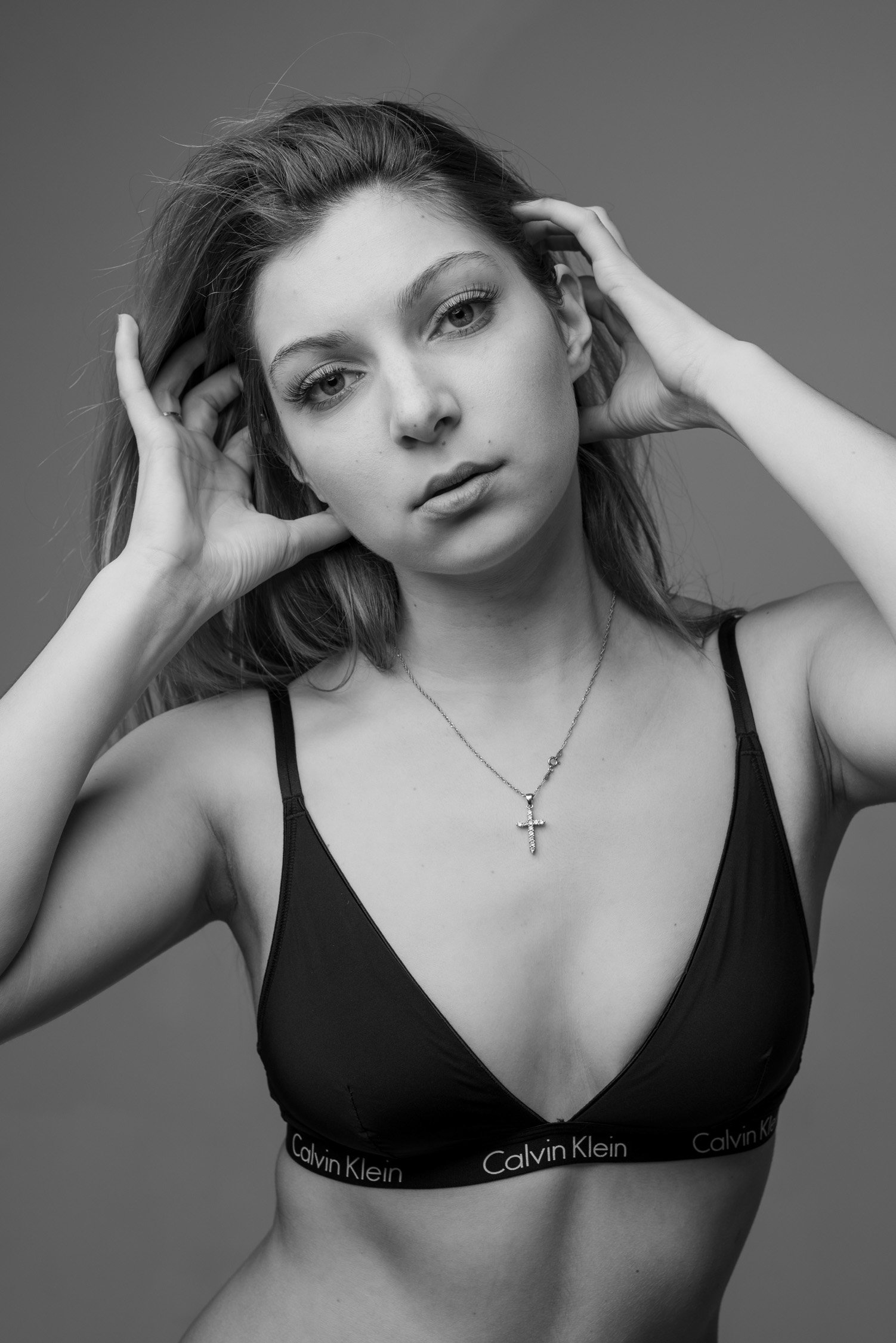 Black and white portrait of a young woman wearing a Calvin Klein sports bra and a necklace with a cross pendant, touching her hair and looking at the camera.
