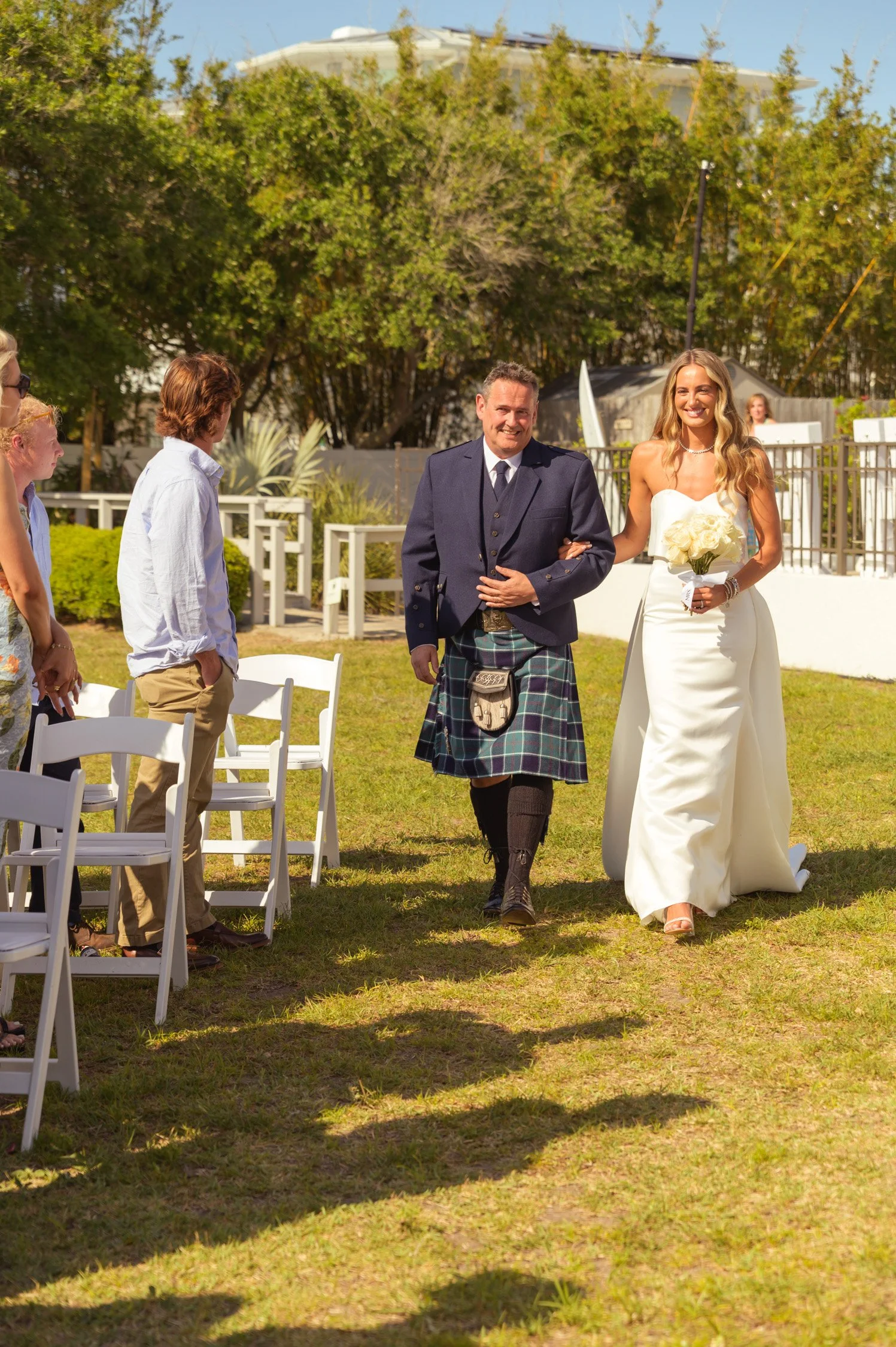 A bride walking down the aisle with a man dressed in formal attire, surrounded by seated guests at an outdoor wedding on a sunny day.