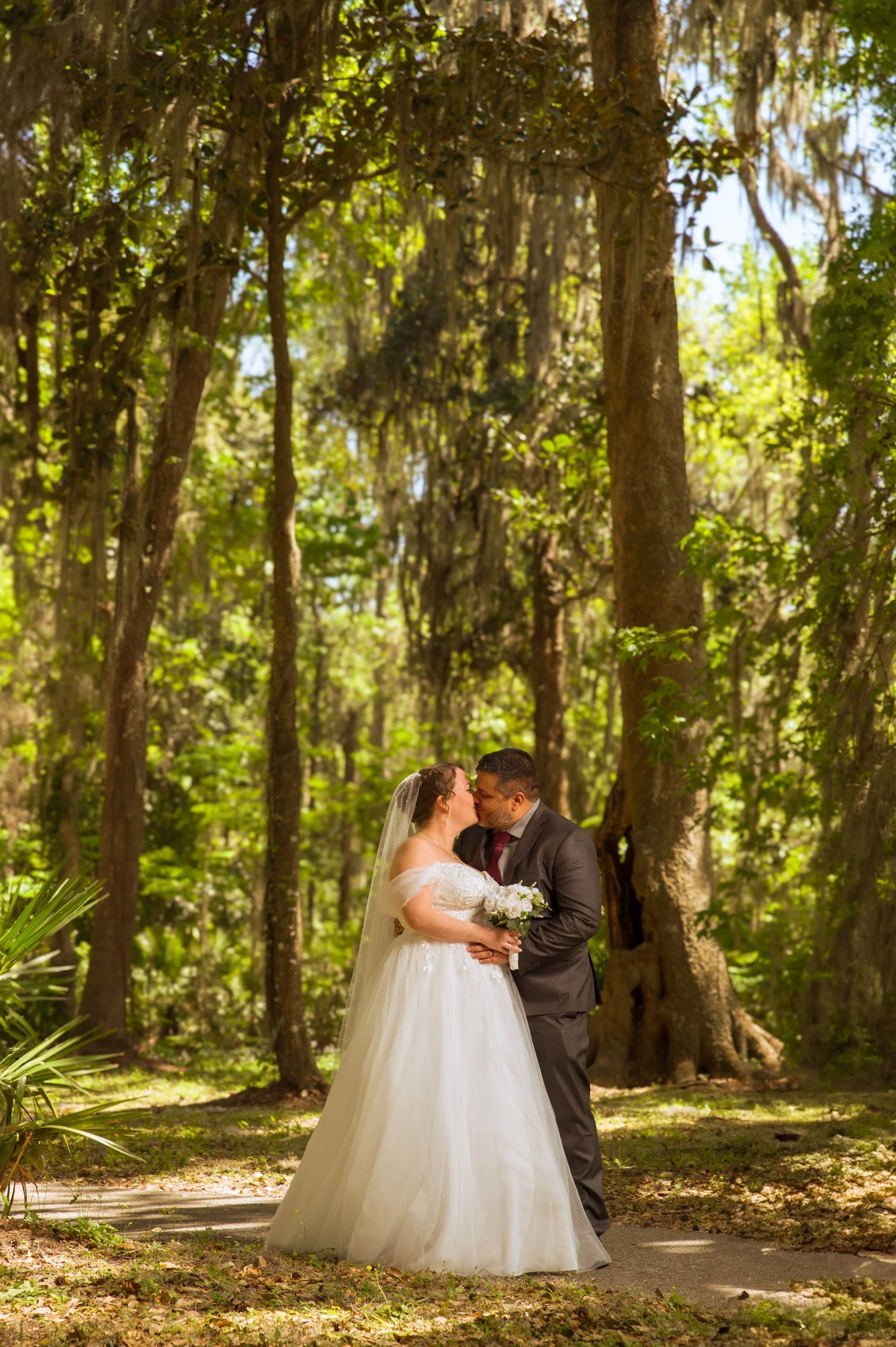A bride and groom sharing a kiss in a forest, with greenery and tall trees surrounding them, during their wedding ceremony.