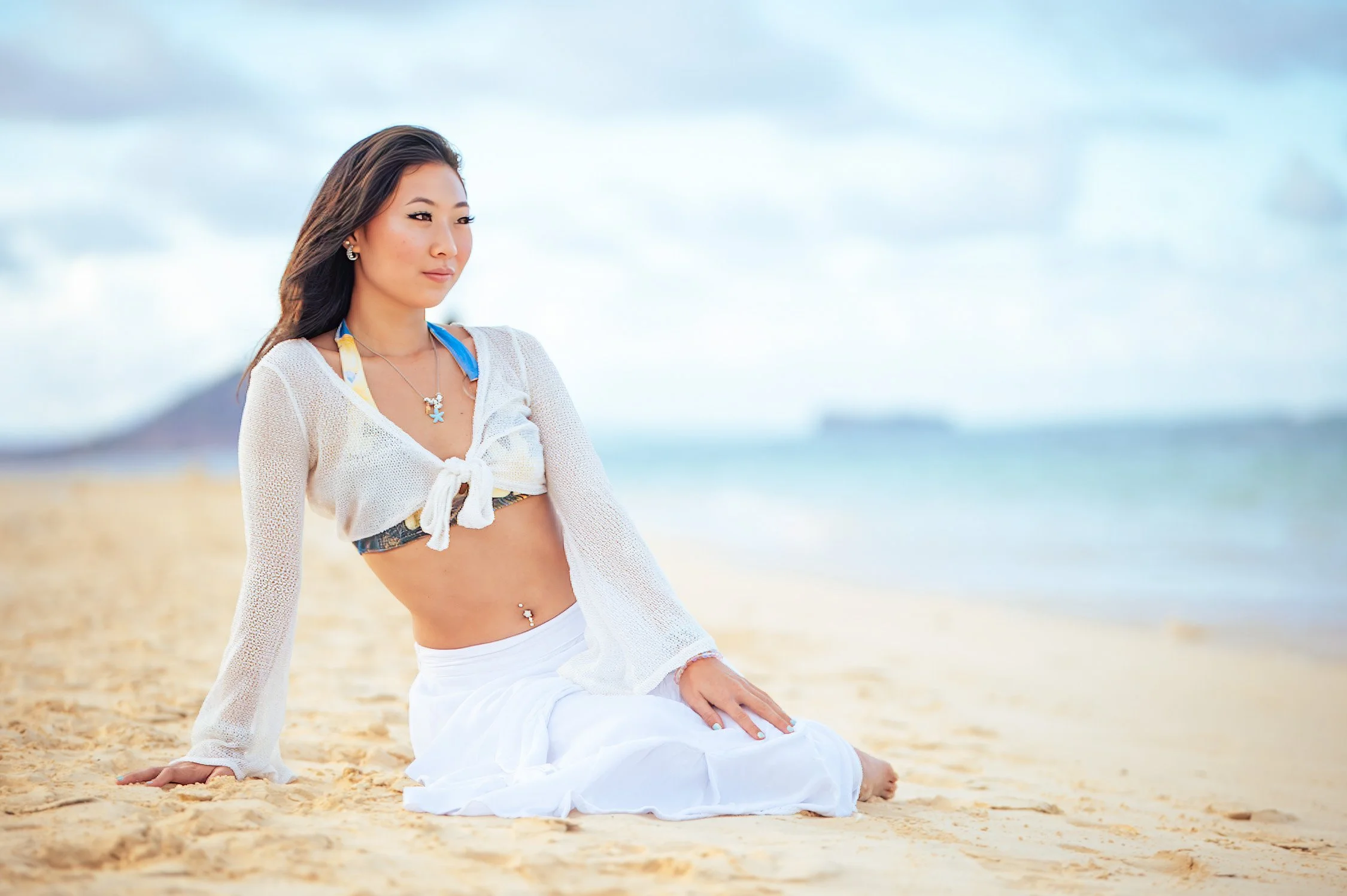 Woman sitting on sandy beach with ocean and cloudy sky in background, wearing a white crochet cover-up, bikini top, white skirt, and jewelry.