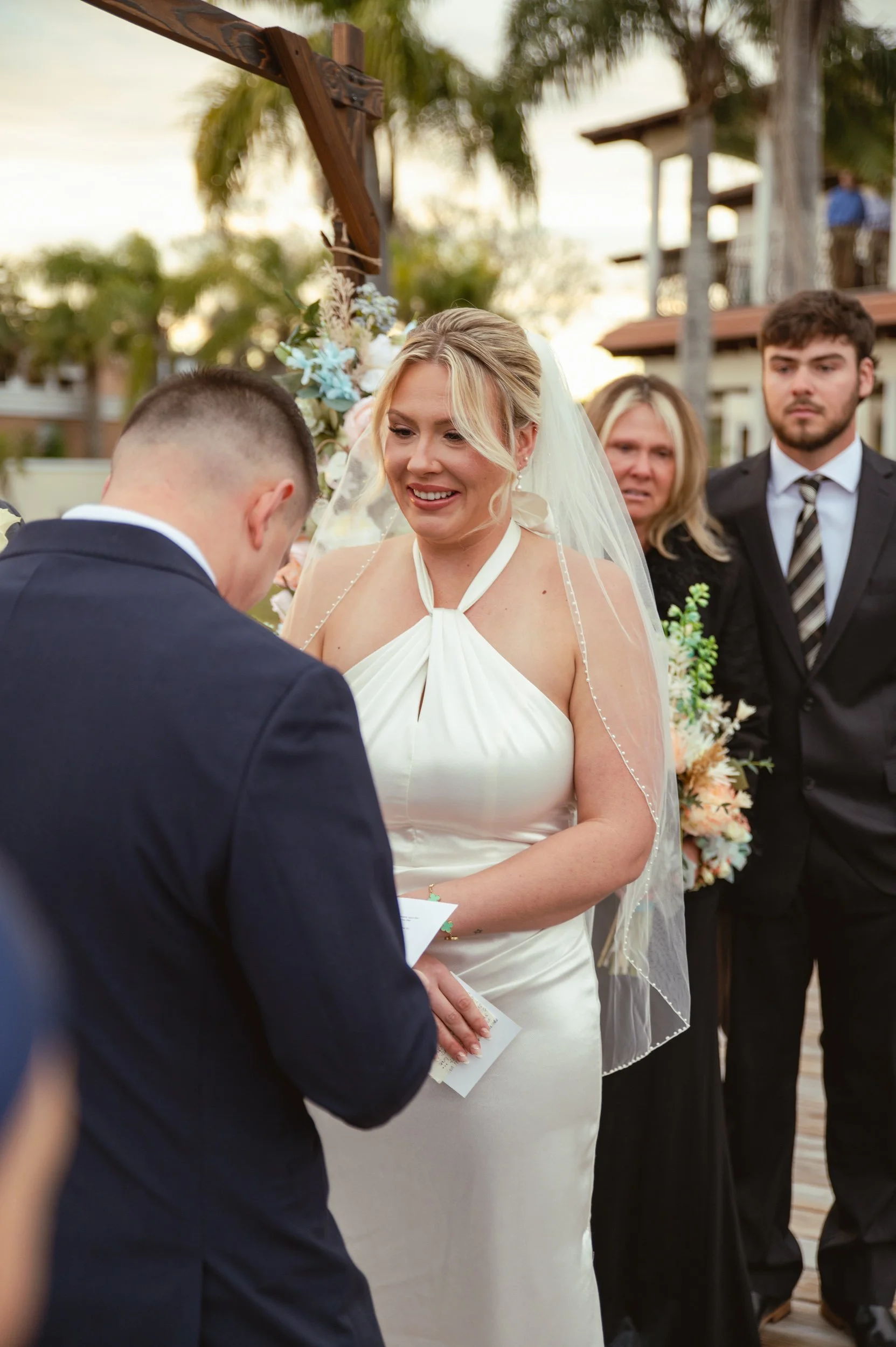 A bride in a white wedding gown exchange vows or rings with a groom during an outdoor wedding ceremony, surrounded by friends and family.